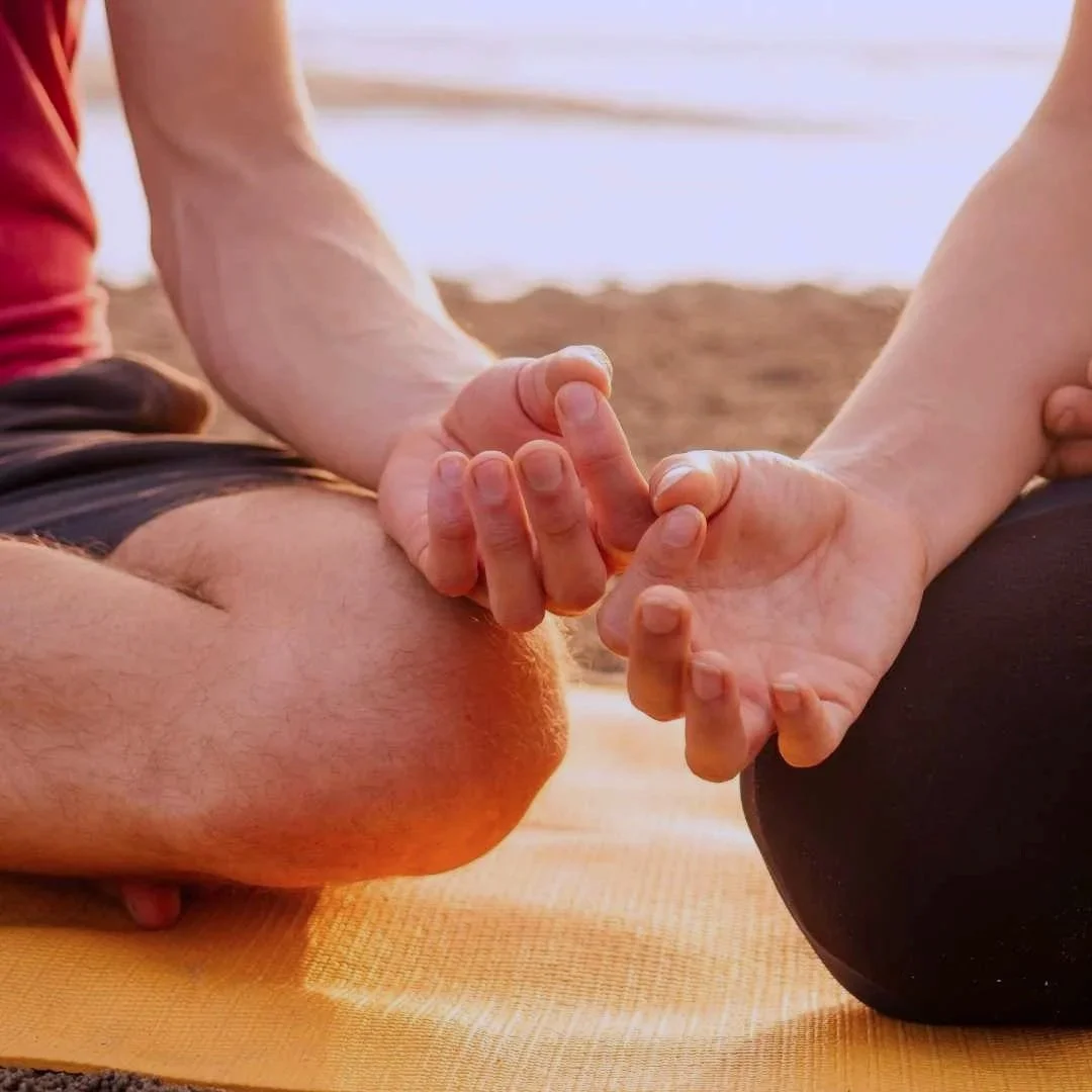 Close-up of two people practicing partner yoga or meditation on a sandy newcastle beach during sunset, holding hands in a mudra gesture while sitting cross-legged.
