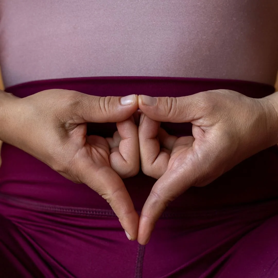 woman hands forming a yoni mudra wearing a pink top and purple pants.
