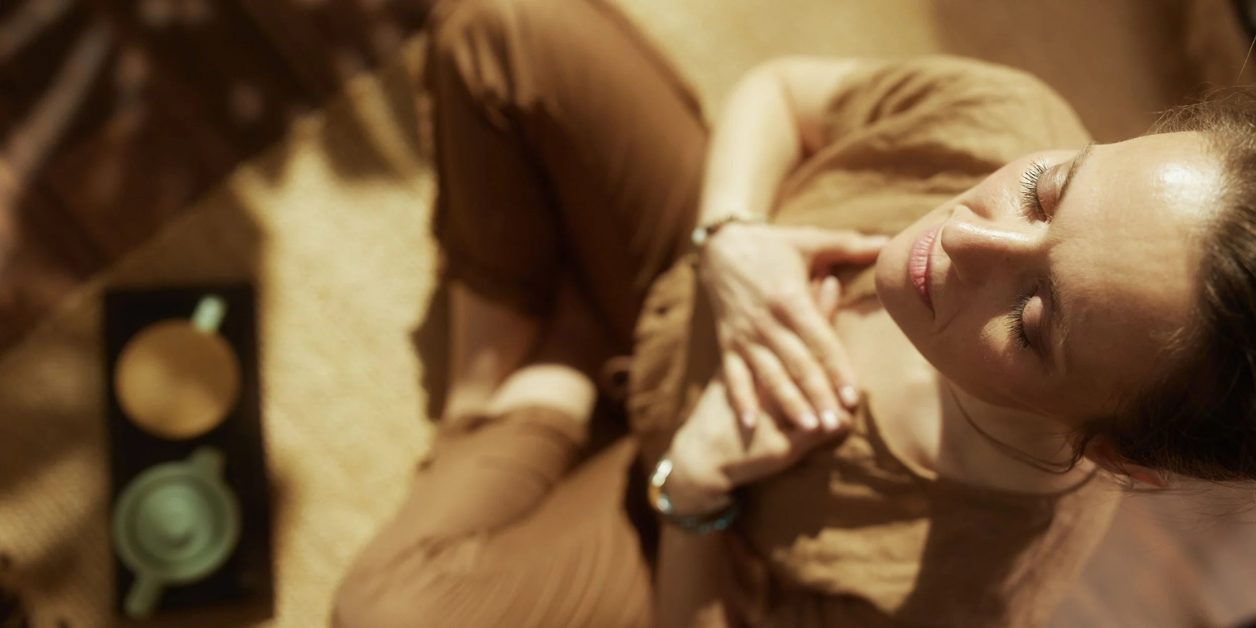 A woman with light skin and dark wavy hair doing tantra breathwork in newcastle with her eyes closed, smiling gently, surrounded by makeup products.