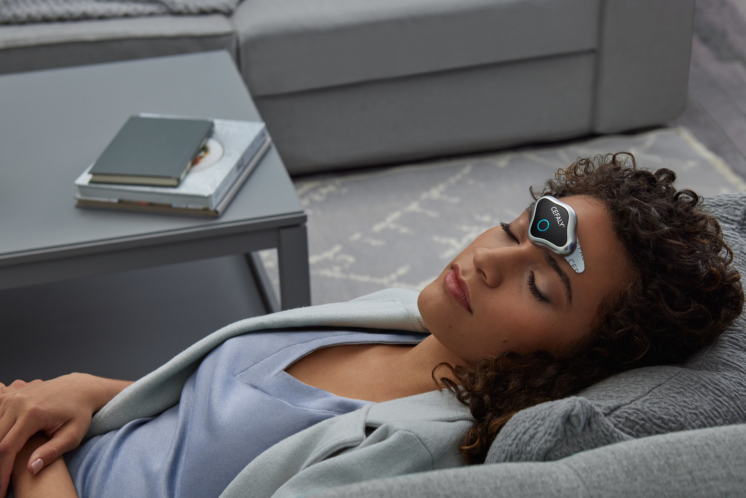 Young woman with curly hair lying on a gray couch with her eyes closed, wearing a wellness device on her forehead labeled 'CEQUALY'.