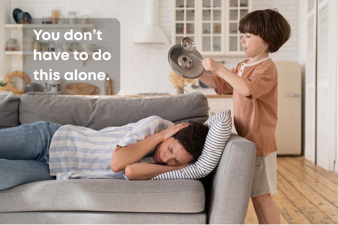 A young boy is standing by a couch and holding a pot lid over a woman lying on the couch, who appears to be asleep or resting with her eyes closed. The boy is smiling and looking at the woman, and there is a speech bubble with the text 'You don’t have to do this alone.'