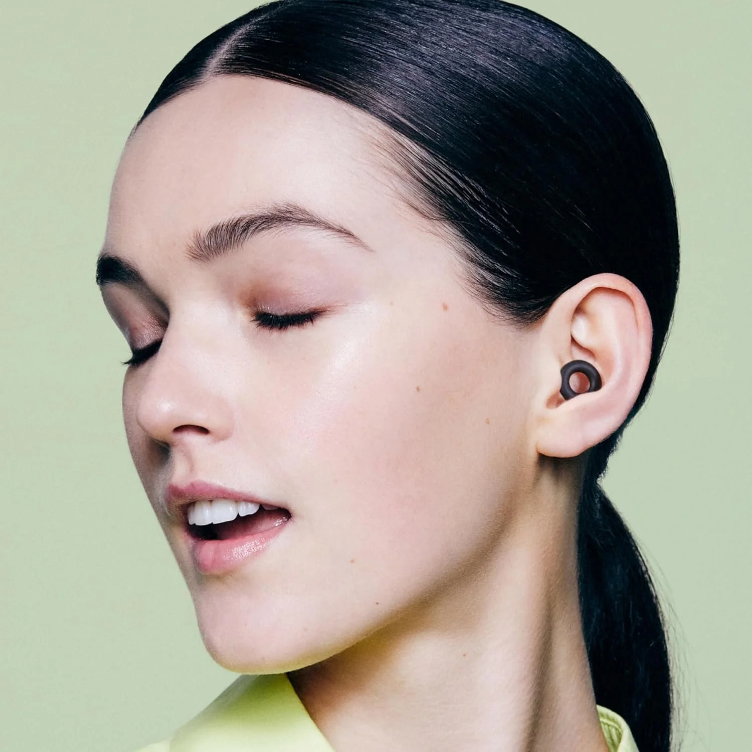 A woman with dark hair styled back, wearing black earrings, with her eyes closed and mouth slightly open, in front of a light green background.