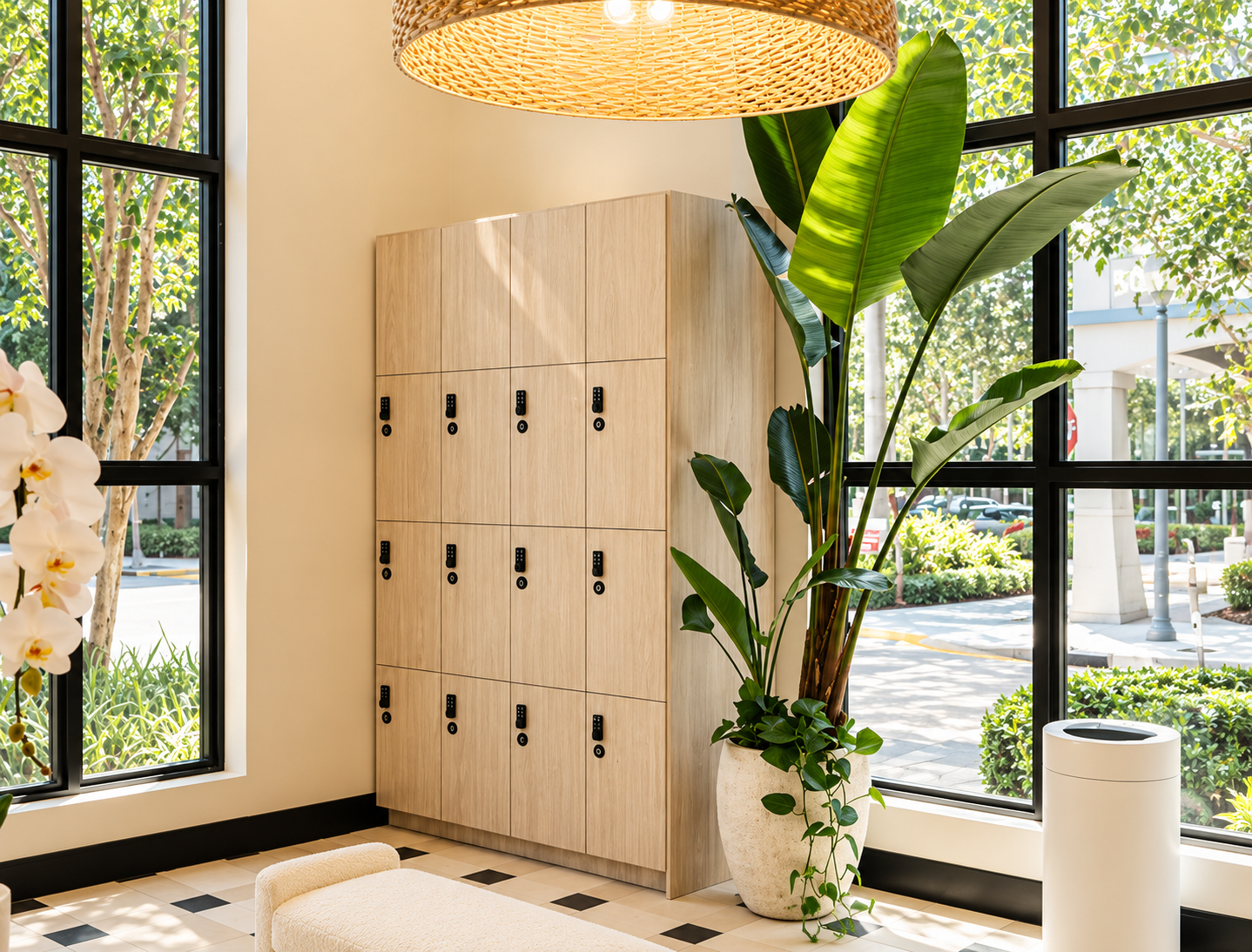 Interior of a modern space with a large window showing greenery outside, a light-colored wooden locker, a large potted plant, a white ottoman, and a woven pendant light hanging from the ceiling.
