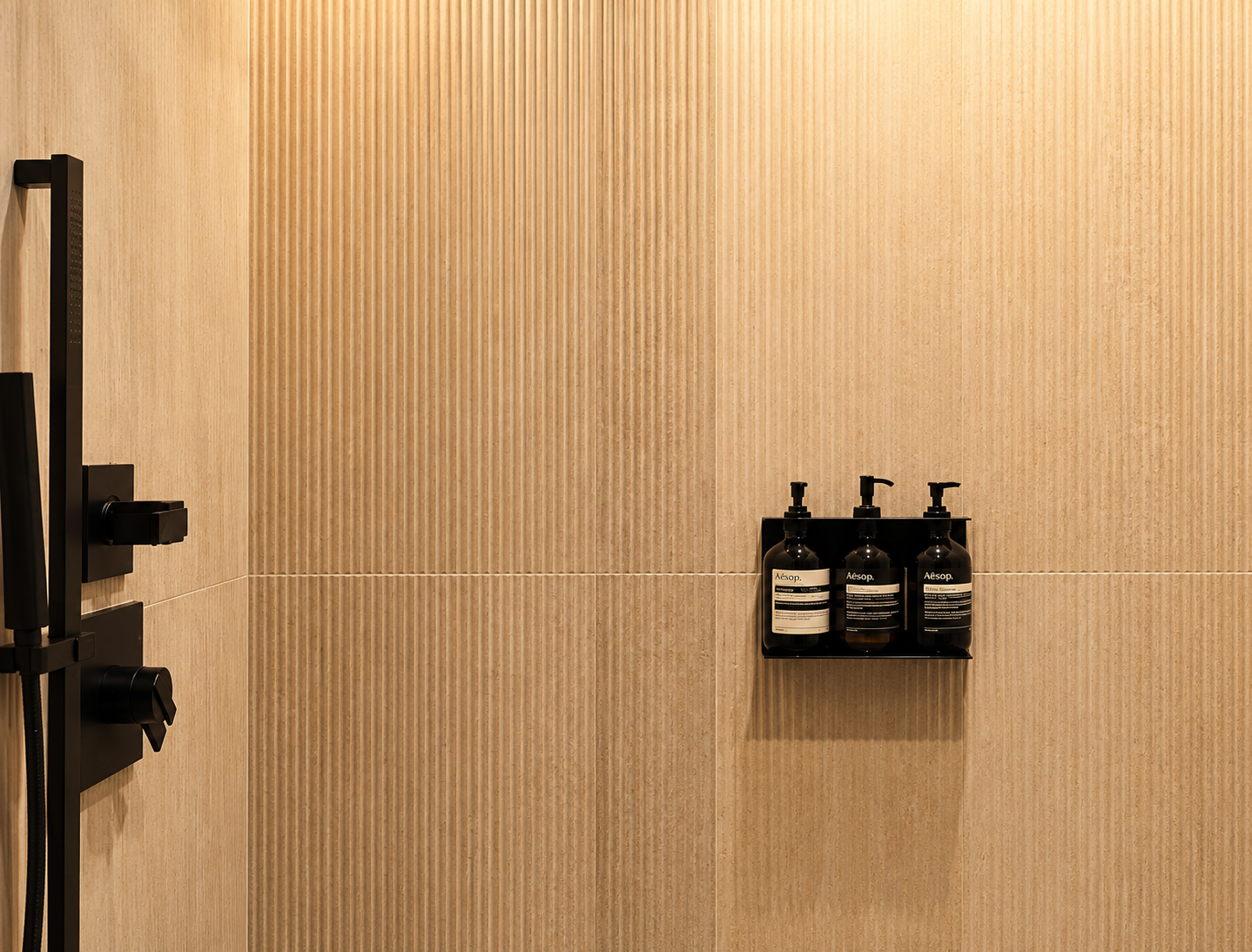 A bathroom shower with beige tiled wall, black shower fixtures, and a wall-mounted shelf holding three black pump bottles of toiletries.