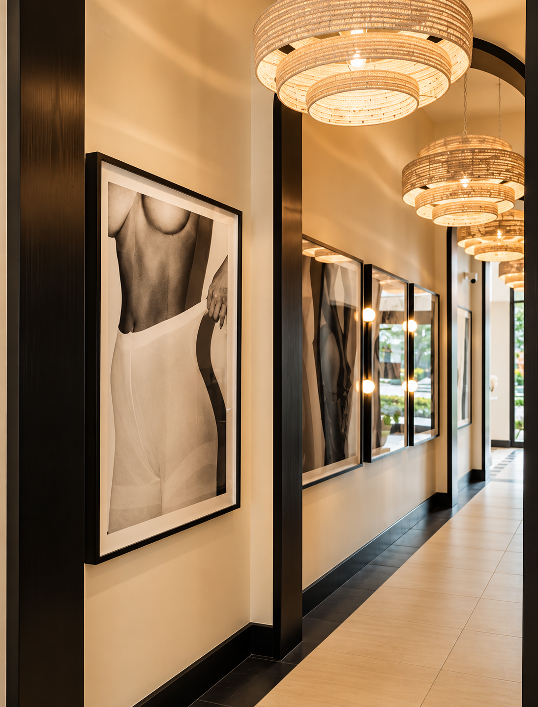 Interior of a modern art gallery with black and white photographs of women's bodies hanging on a cream-colored wall, framed in black. The ceiling has multiple round woven pendant lights, and the floor is tiled with black and light beige sections. Lar