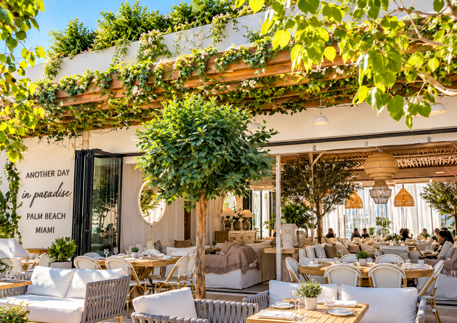 An outdoor restaurant with white and beige seating, wooden tables, potted plants, decorative lighting, and greenery including trees and vine-covered pergola, set against a clear blue sky.