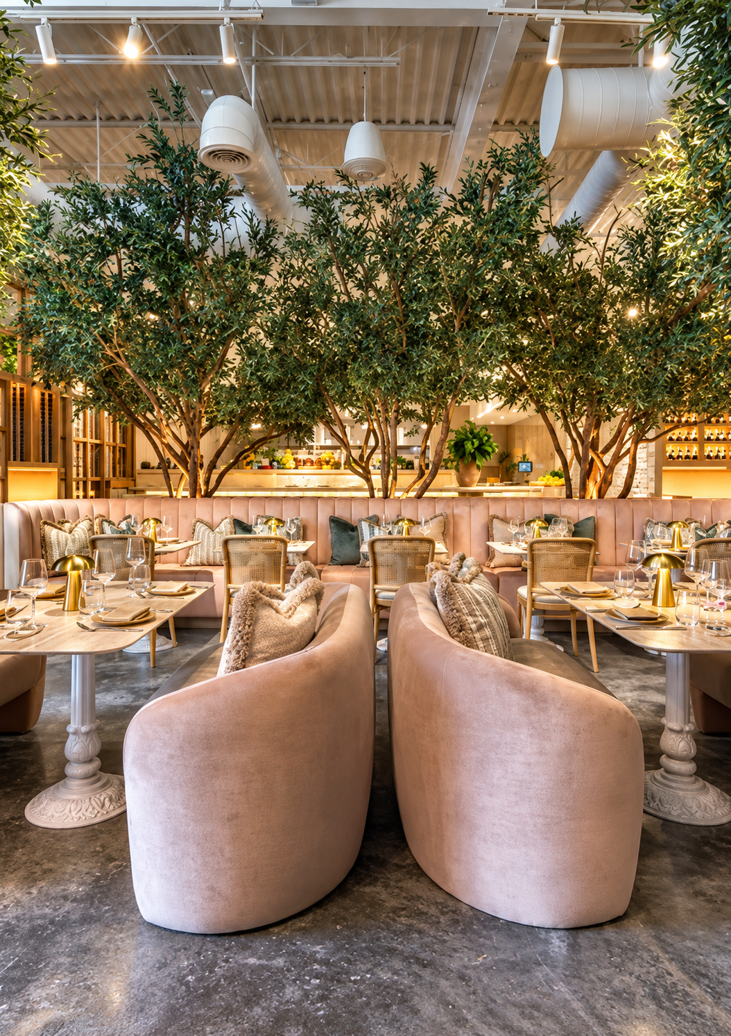 Elegant restaurant interior with pink velvet booths, wooden tables, and chairs, surrounded by lush green trees and decorative pillows, with warm lighting and a modern, cozy ambiance.