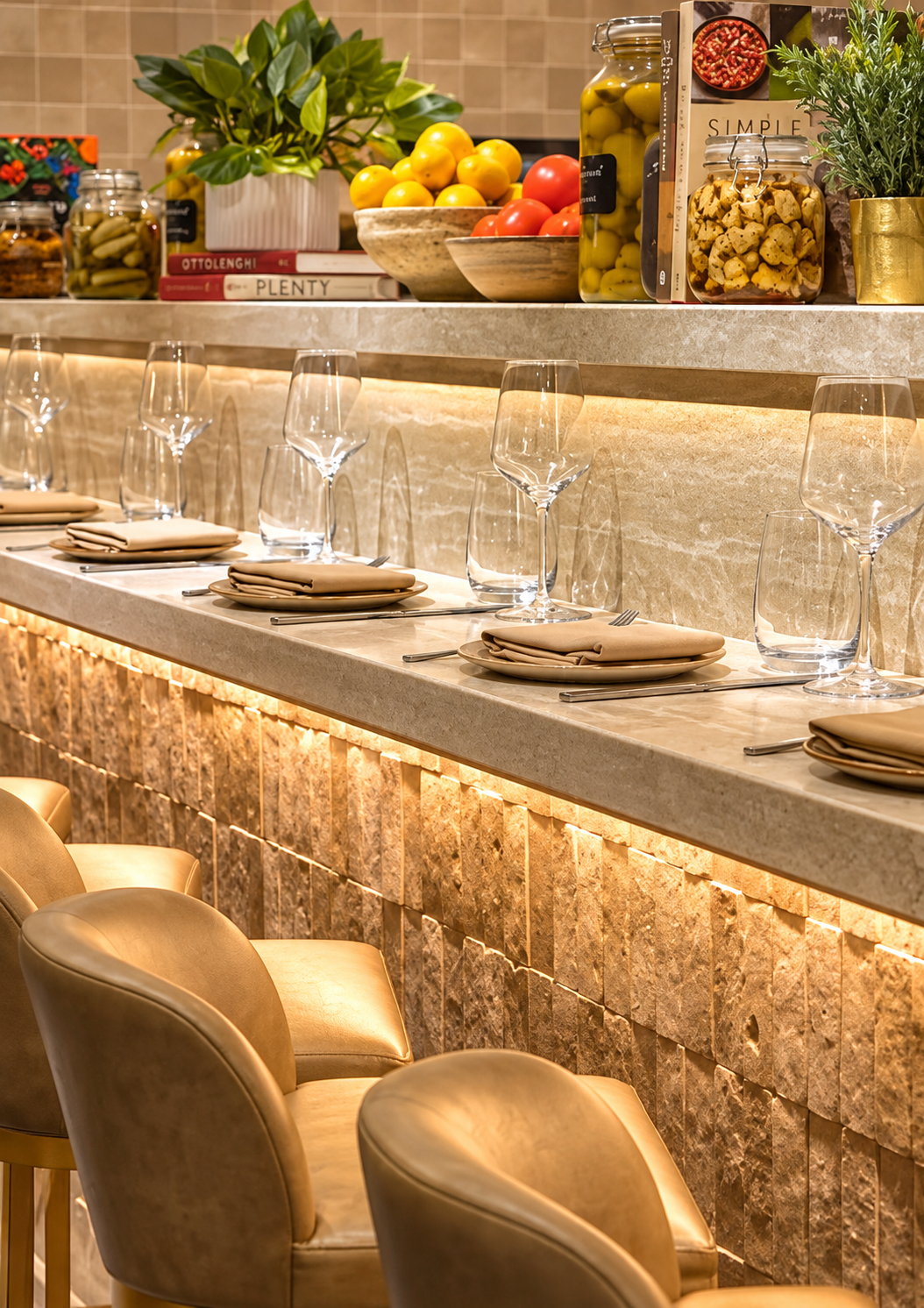 A restaurant bar area with a beige stone countertop, set with gold-colored chairs, beige napkins, plates, and utensils. Behind the bar, there are jars of pickles, a bowl of tomatoes, and jars of olives and artichokes, along with potted plants and boo