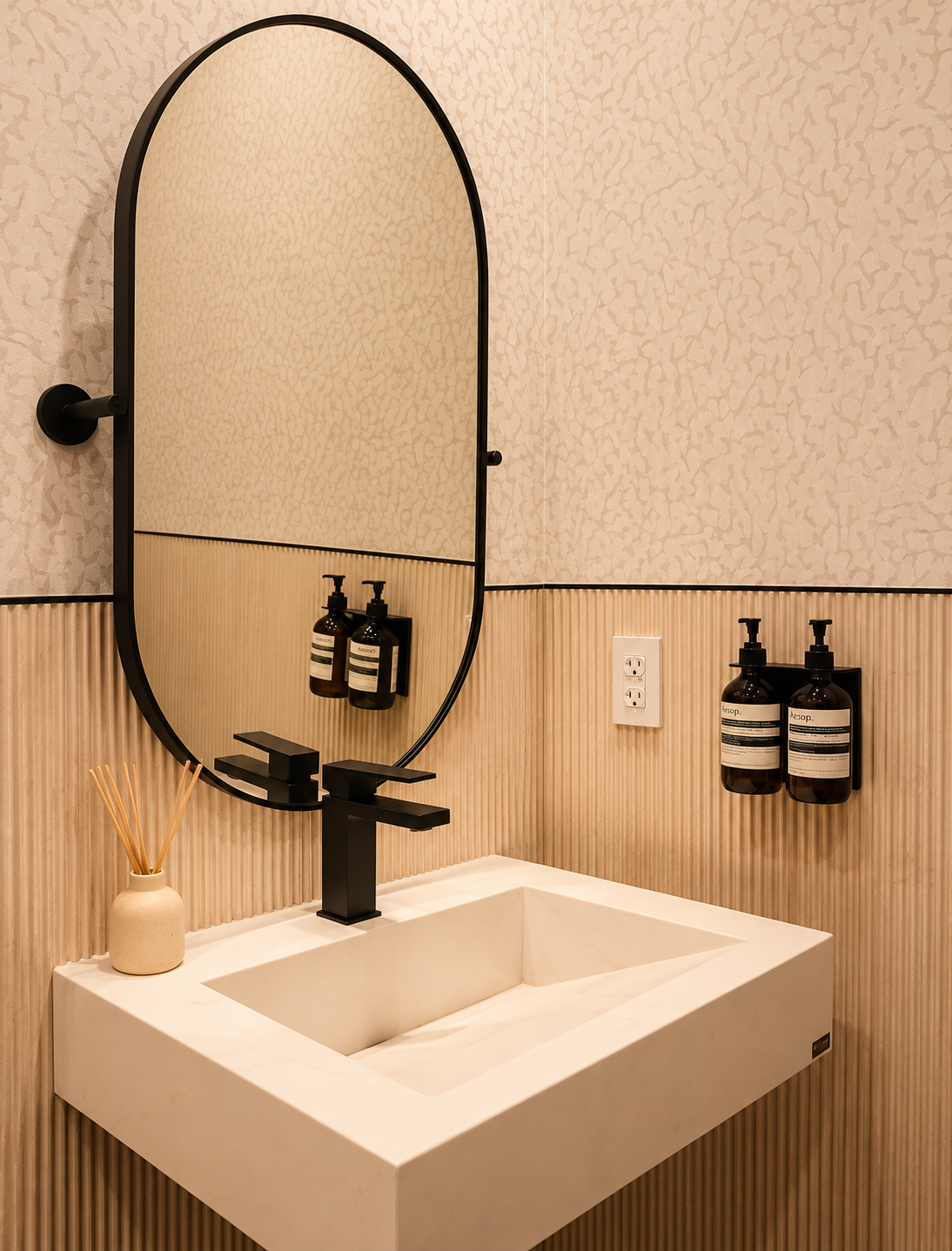 Modern bathroom sink with a black faucet, oval mirror, and wall-mounted soap dispensers, with a decorative vase containing reed diffusers on the sink counter.