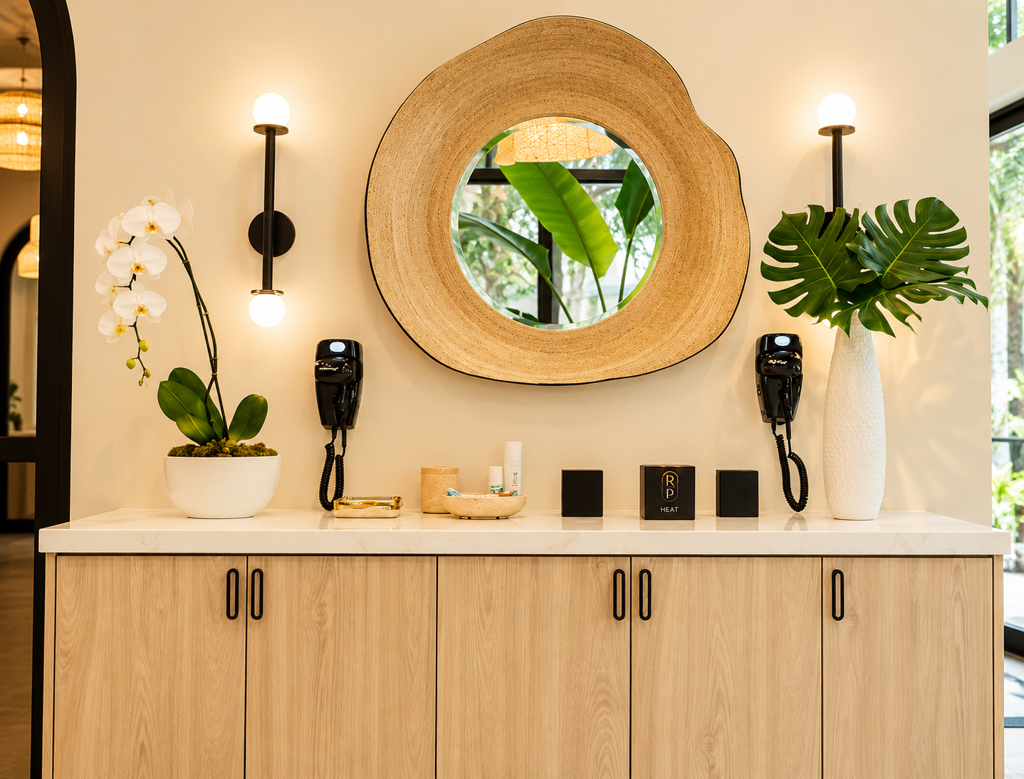 Modern interior wall decor with a large round wooden-framed mirror, two black wall-mounted hair dryers, two potted plants in white vases, a white marble-topped wooden cabinet, and various small objects.