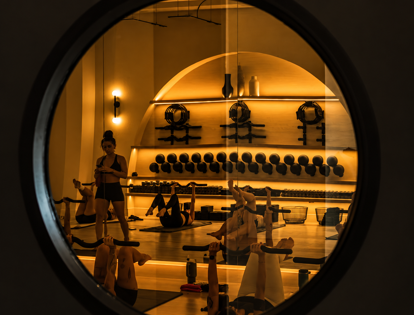A fitness studio with people participating in a workout class, viewed through a circular mirror. The studio has warm lighting, a curved wall with shelves holding weights and equipment, and participants lying on mats doing leg raises or stretches.