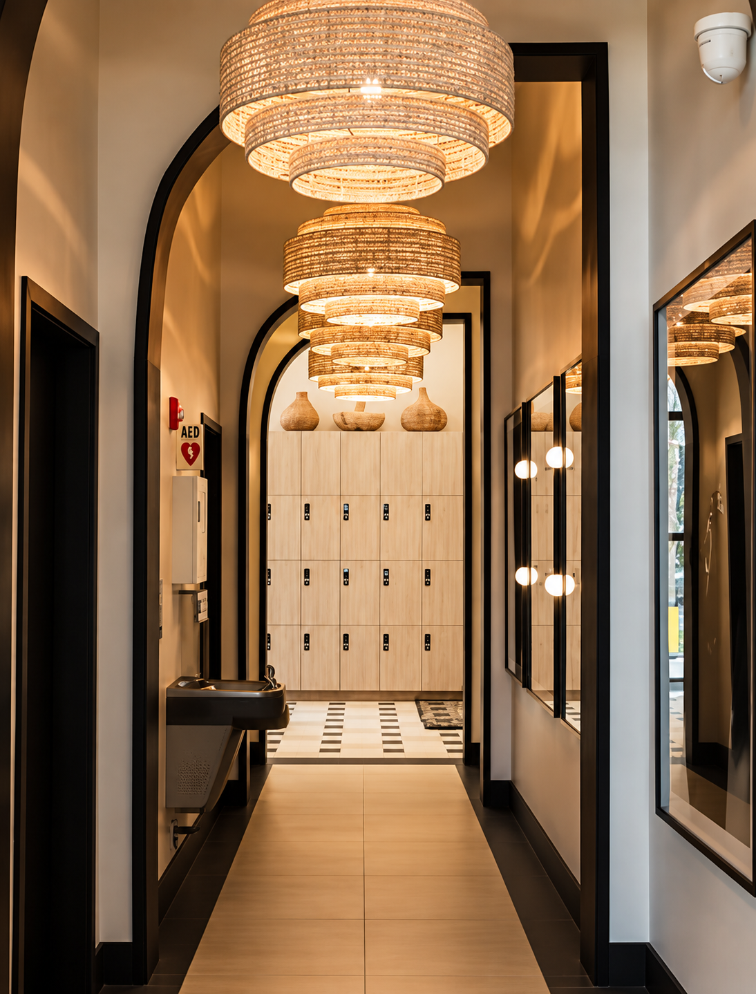 A hallway with modern decor and lighting, wooden lockers at the end, decorative vases on top, and black-framed mirrors on the side walls.