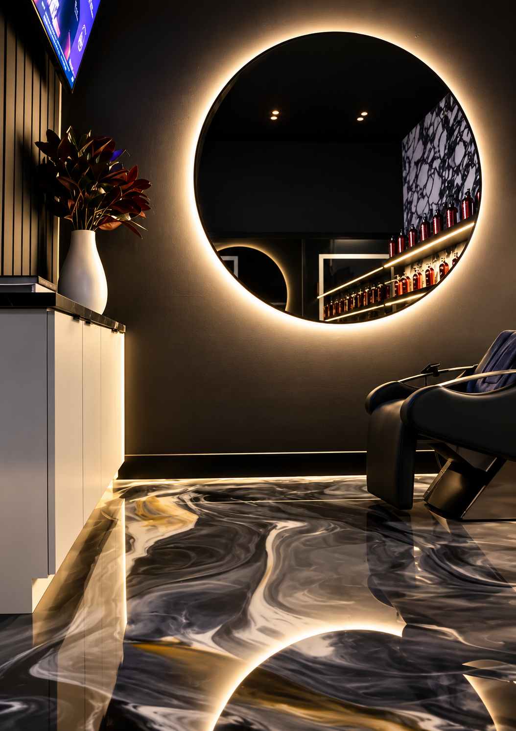 Modern interior room with a large round mirror backlit with warm light, a black salon chair, a white cabinet with a decorative vase of dark red foliage, and a marble-patterned floor with swirling black, white, and gold design.