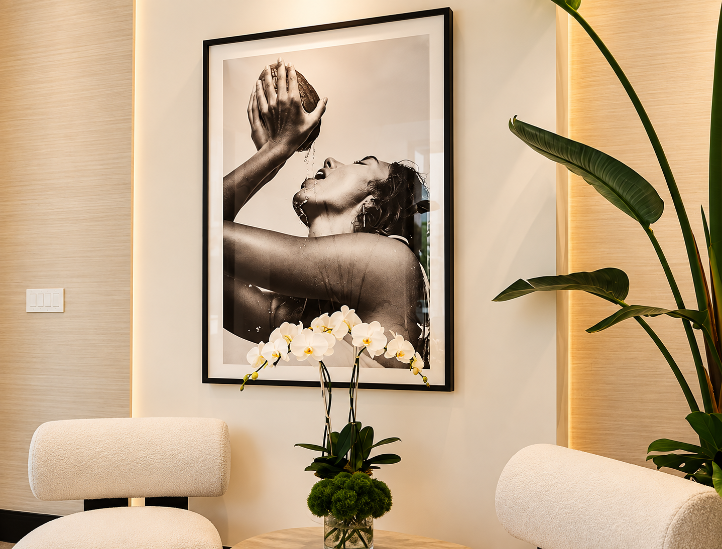 Decorative room with a framed black and white photograph of a woman holding an apple, potted orchids on a table, leafy houseplants, and cream-colored chairs.
