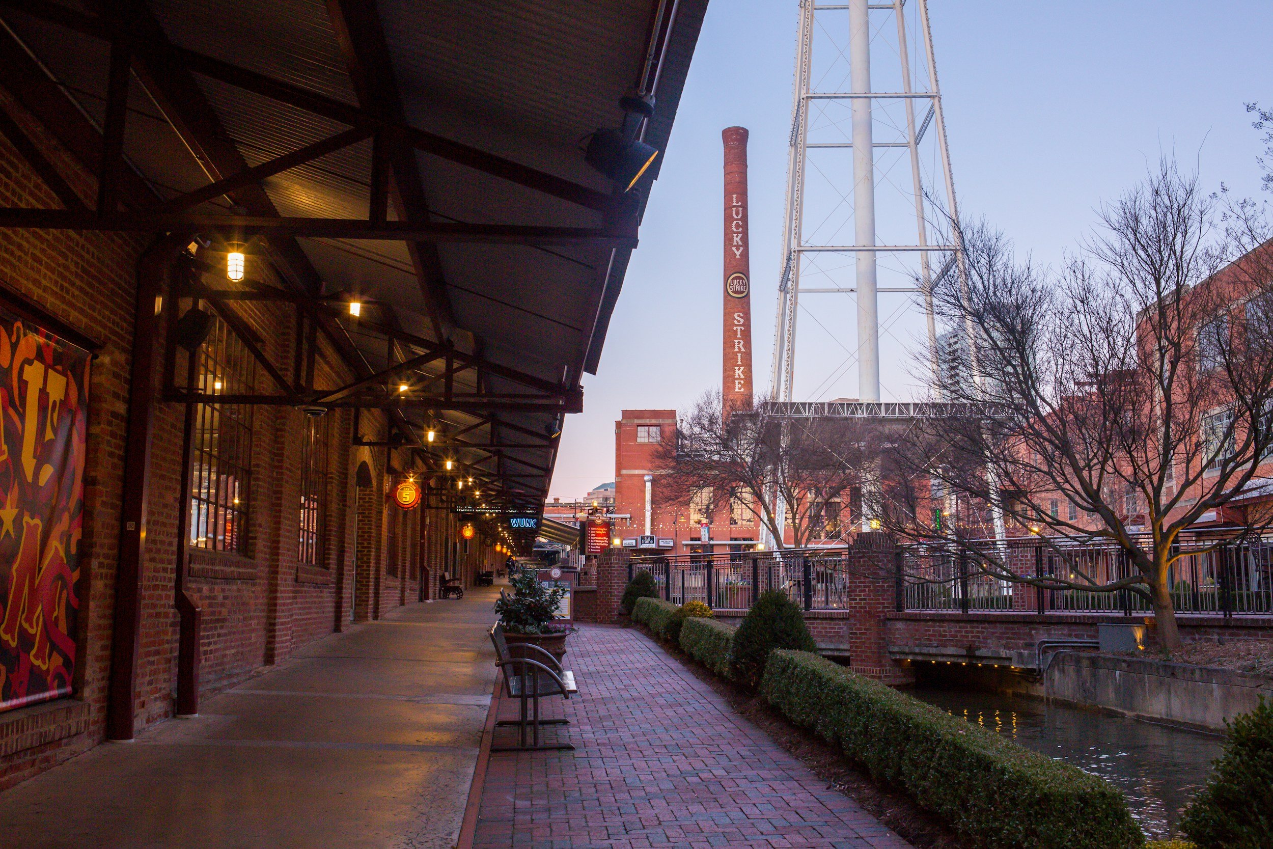 View of a brick sidewalk along a waterway with benches, small bushes, and a metal railing. Building with string lights and large windows on the left. Tall radio tower, smokestack marked 'Lucky Strike' and leafless trees in the background against a clear sky.