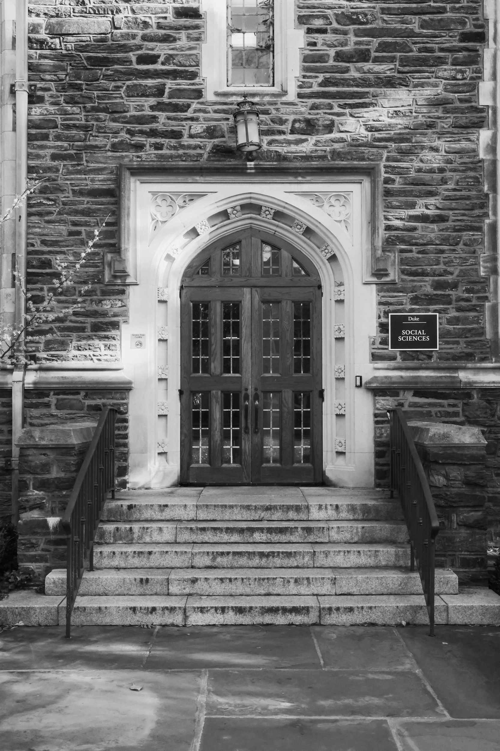 The image shows the entrance to a stone building with steps leading up to a wooden double door. There is ornate trim around the door, a lantern-style light fixture hanging above, and a small window above the door. To the right of the door, there is a sign that reads 'Duke Social Sciences.'