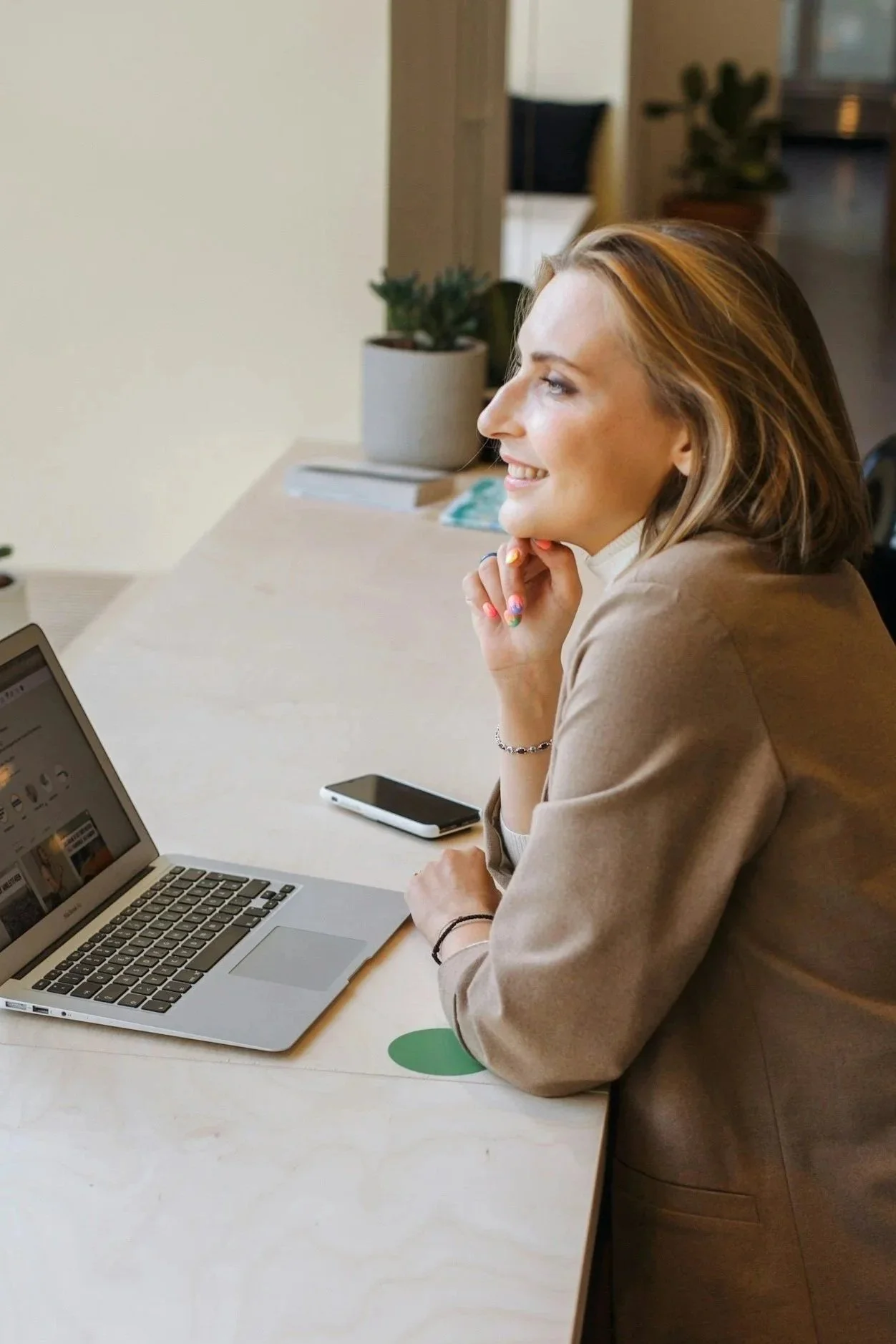 A woman with blonde hair sitting at a light-colored wooden table with an open laptop in front of her, smiling and looking to her right. There is a smartphone next to the laptop, and potted plants in the background.