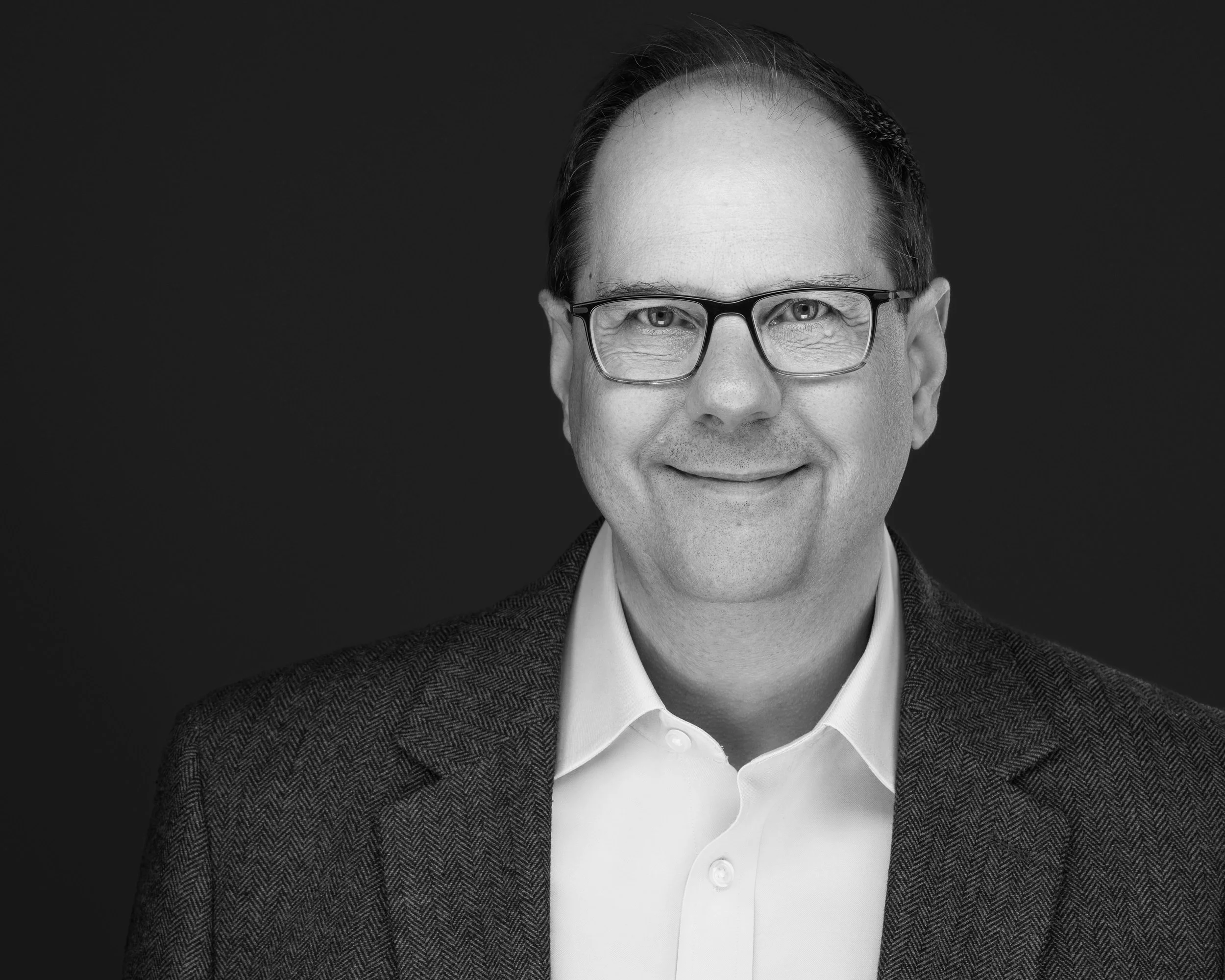 Black and white portrait of a smiling man wearing glasses, a white shirt, and a dark blazer against a plain background.