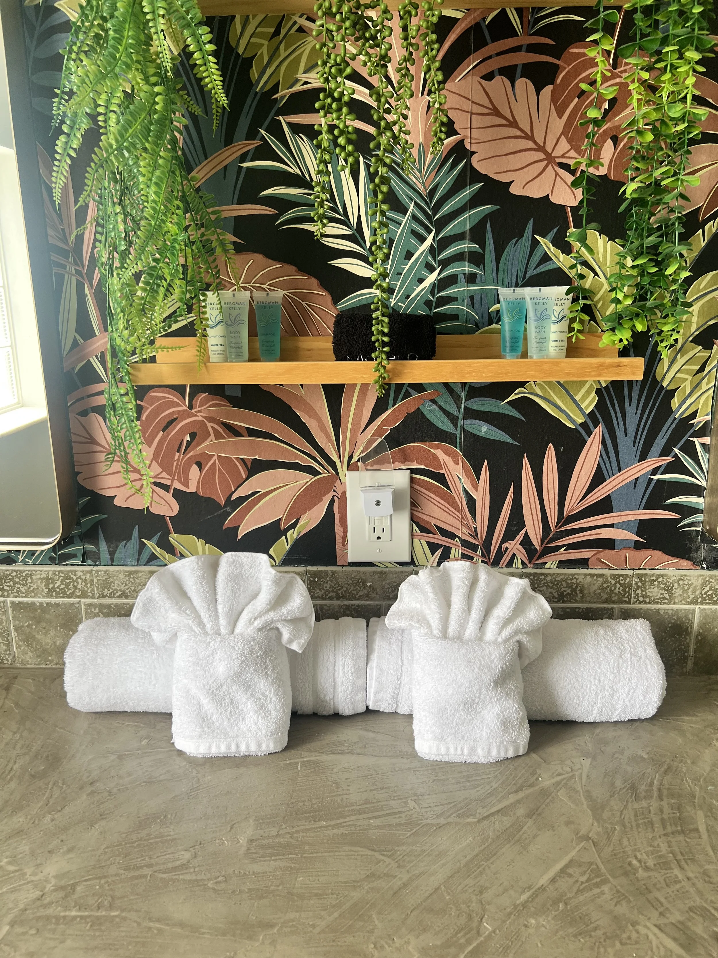 Bathroom decor featuring a wooden shelf with various toiletries, lush green hanging plants, and a background with tropical leaf mural. Two towel sculptures resembling flowers made from white towels are placed on the floor beneath the shelf.