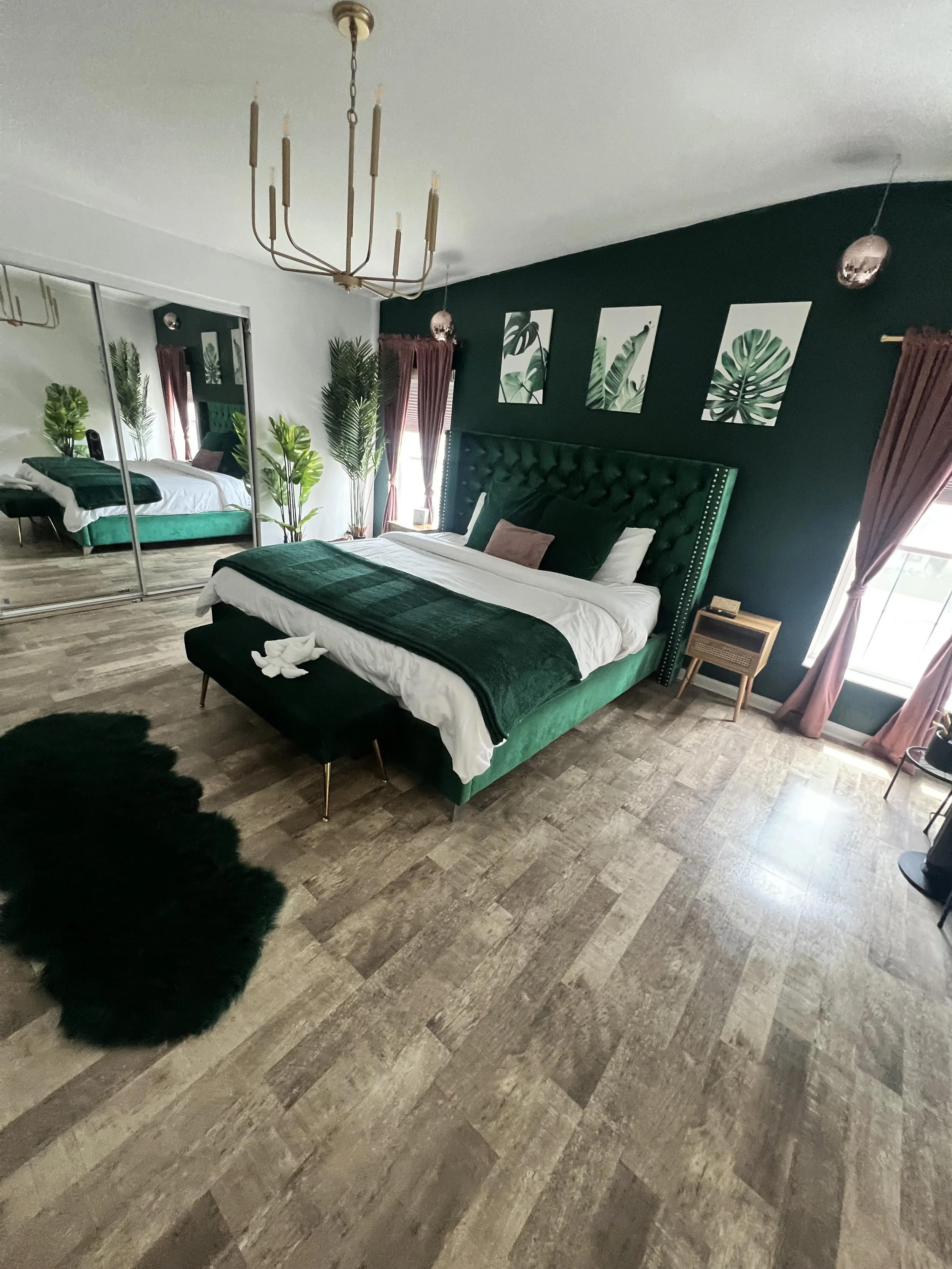 Bedroom with a large green upholstered bed, dark green accent wall with three botanical art prints, mirrored closet, and multiple indoor plants, including a potted palm, with pink curtains and a light wood floor.