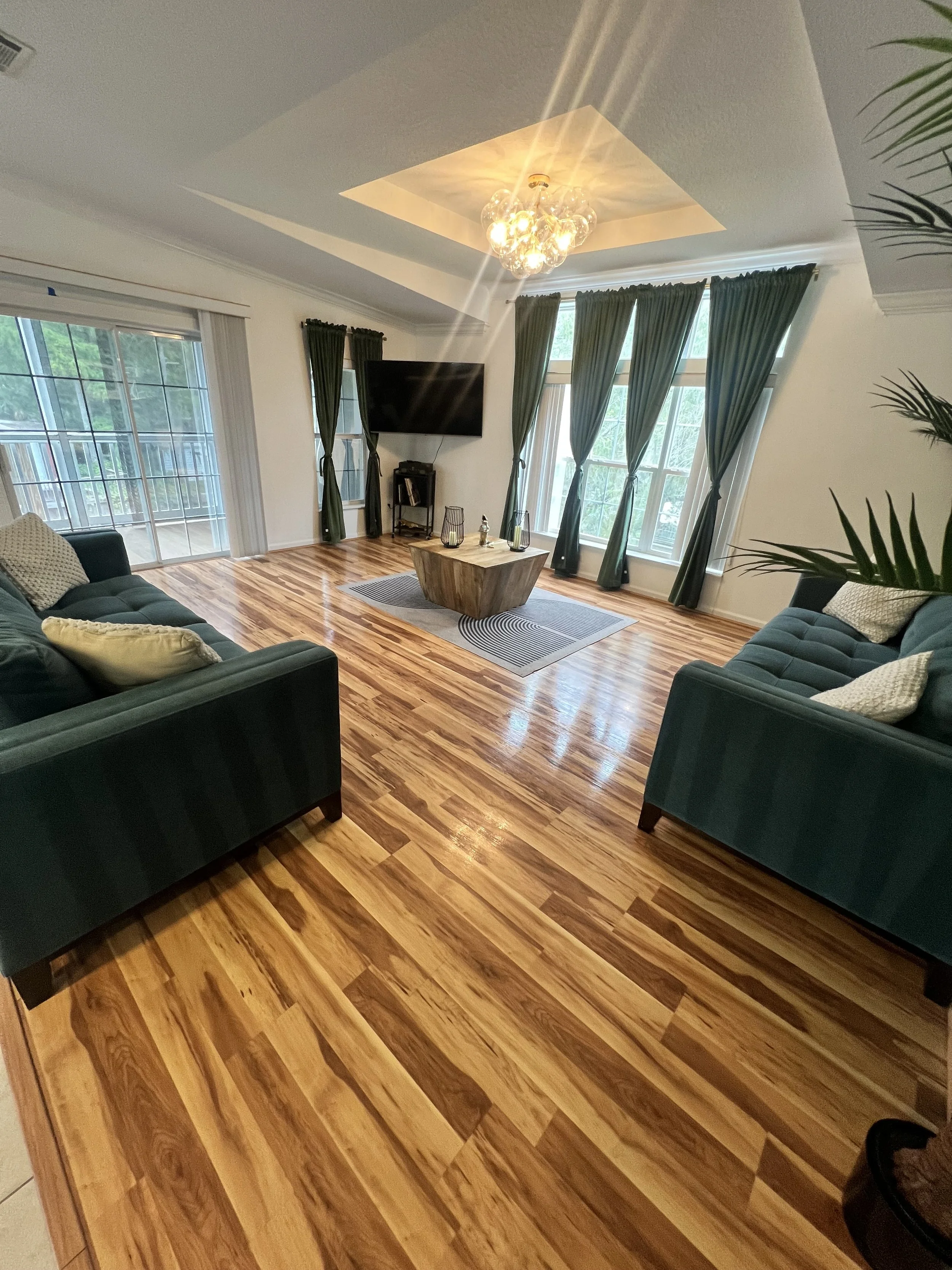 Living room with hardwood floor, two green sofas with beige pillows, a wooden coffee table, a wall-mounted flat-screen TV, large windows with dark green curtains, and a chandelier hanging from a tray ceiling.