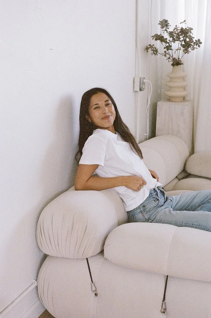 A young woman with long dark hair wearing a white t-shirt and jeans sitting on a light-colored sofa, smiling with a slight tilt of her head, in a bright, minimalistic room with a white wall, a potted plant, and sheer curtains.