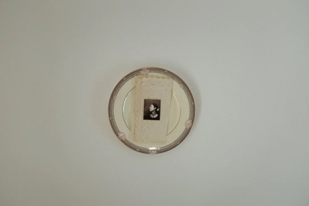 Close-up of a round decorative mirror reflecting a framed black and white photo of a woman with her hair in a bun, against a beige textured wall.