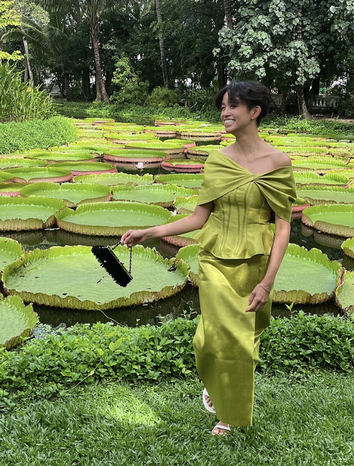 Woman in a shiny green dress standing next to a pond with large green lily pads, holding a small black purse, smiling amid lush greenery and trees.