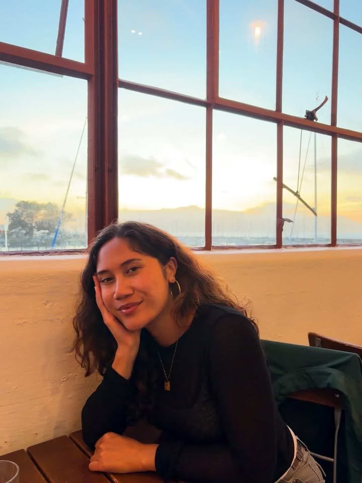 A young woman with curly hair, wearing a black top, sitting at a wooden table inside a restaurant, with a large window behind her showing a sunset over a body of water.