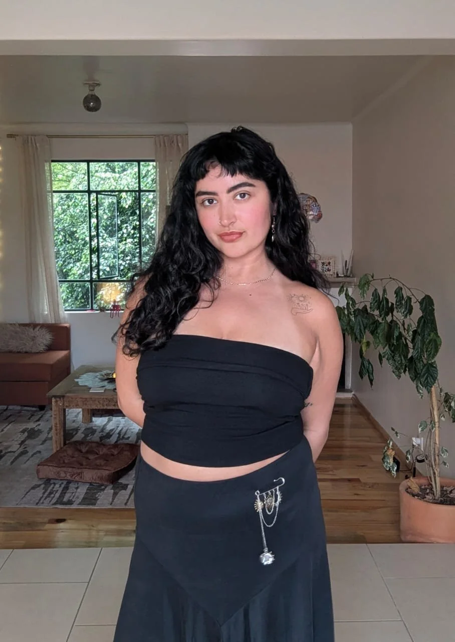 Young woman with curly black hair wearing a black strapless top and black skirt with chain accessories, standing indoors with a window and houseplants in the background.