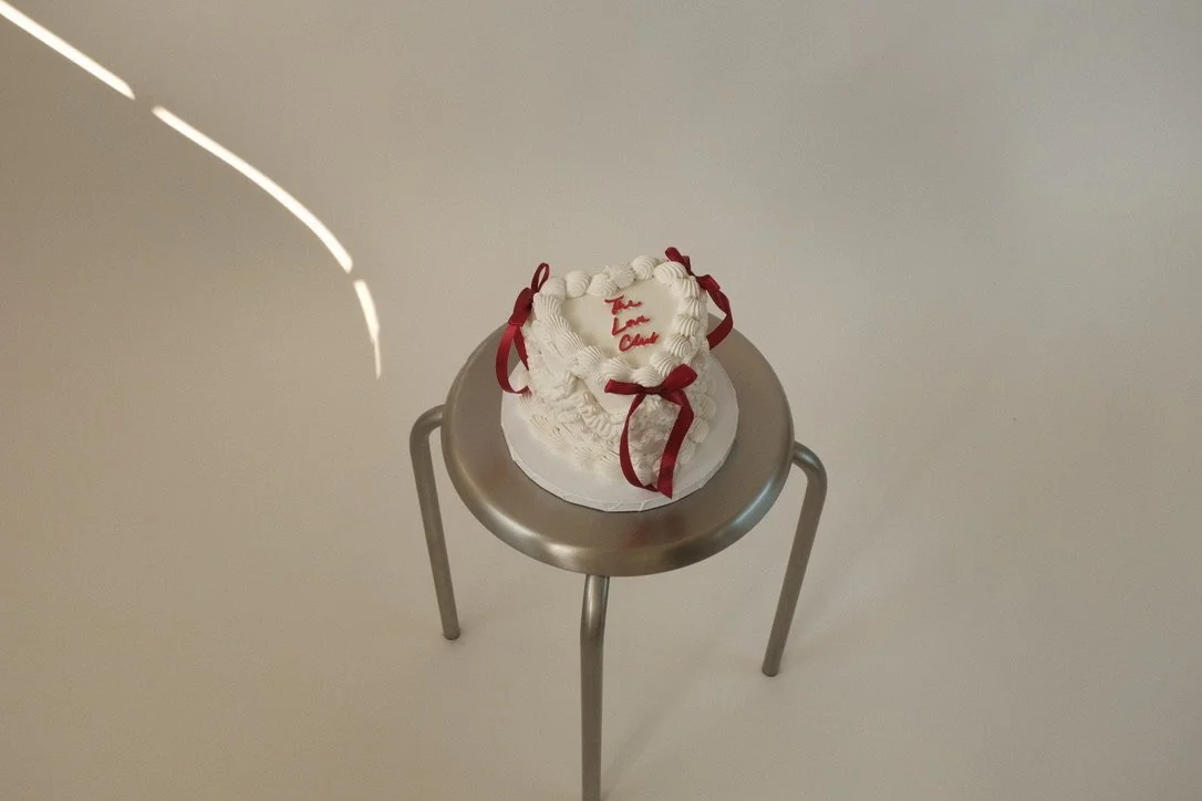 A white cake with red ribbon decorations and the message 'To Love Cared' written on top, placed on a small round silver table.