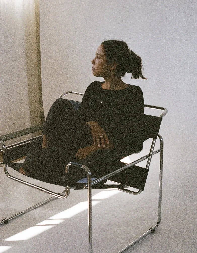 A woman sitting on a modern black lounge chair, looking to her right, with a white wall background.