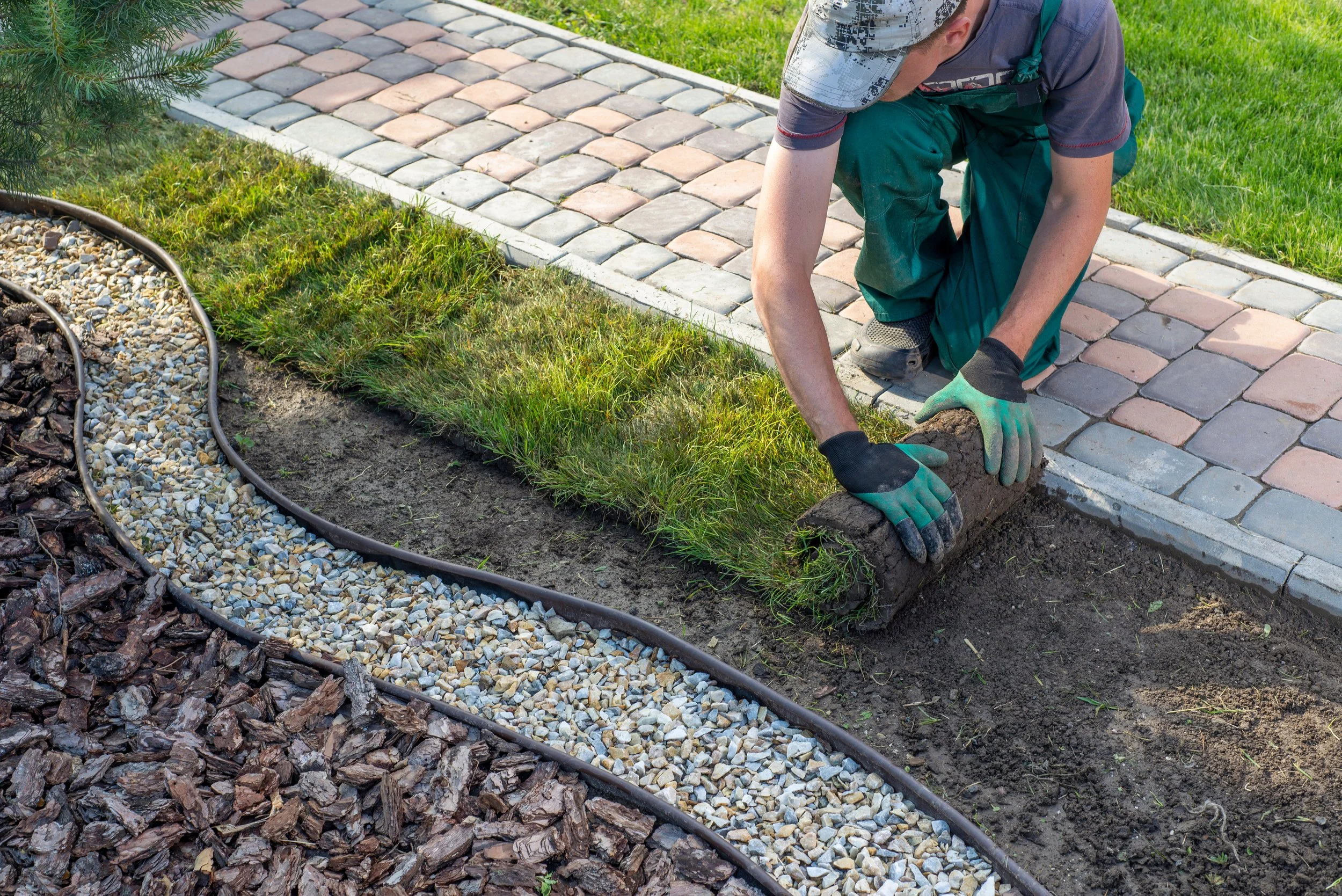 A person kneeling on a stone pathway, laying down pavers to create a decorative border. The individual is wearing a baseball cap, a t-shirt, green work gloves, and green work pants. The scene is outdoors with grass and a landscaped area with mulch and a garden bed nearby.