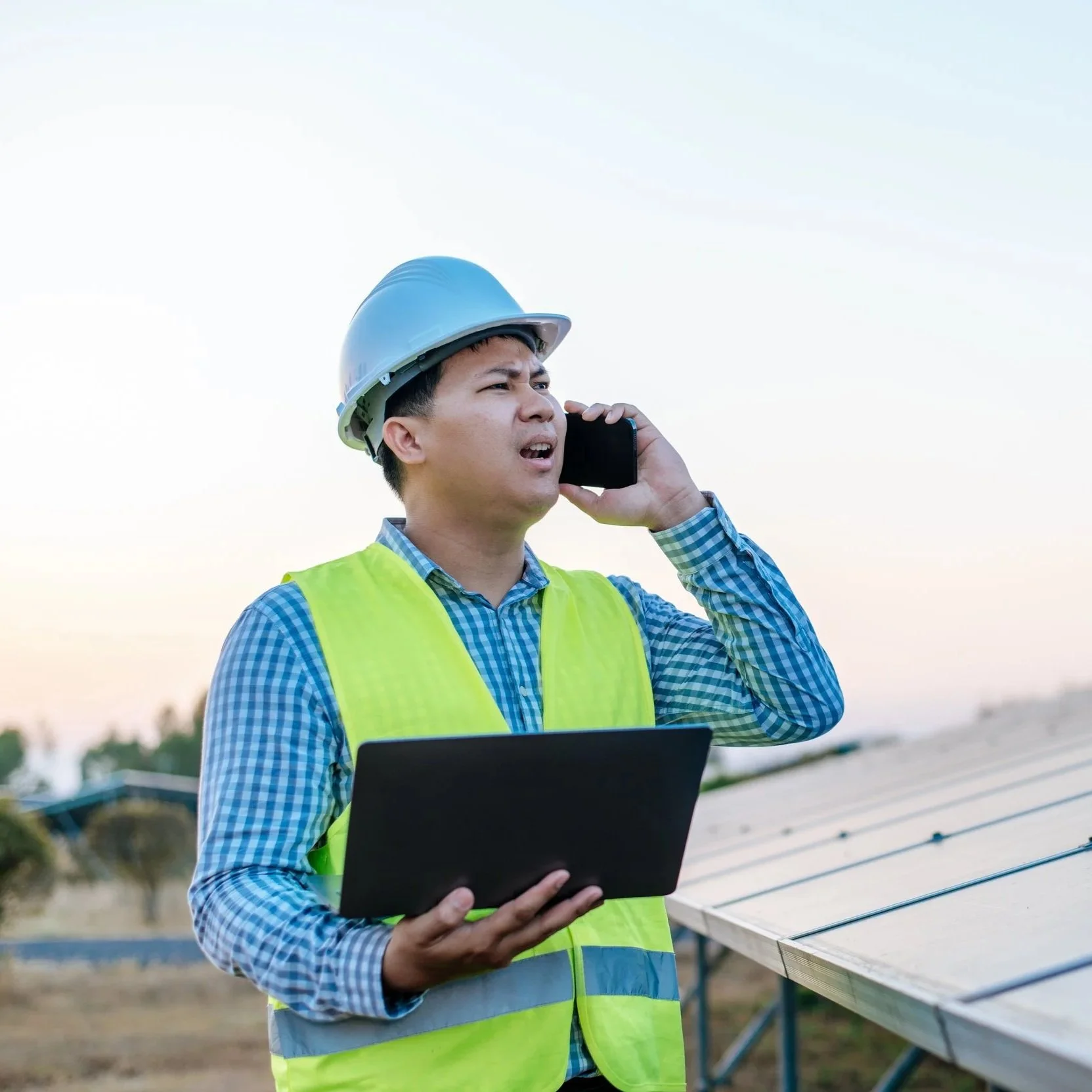 A man wearing a blue checkered shirt, a yellow safety vest, and a white hard hat is standing outdoors near solar panels, talking on a cell phone, and holding a tablet.