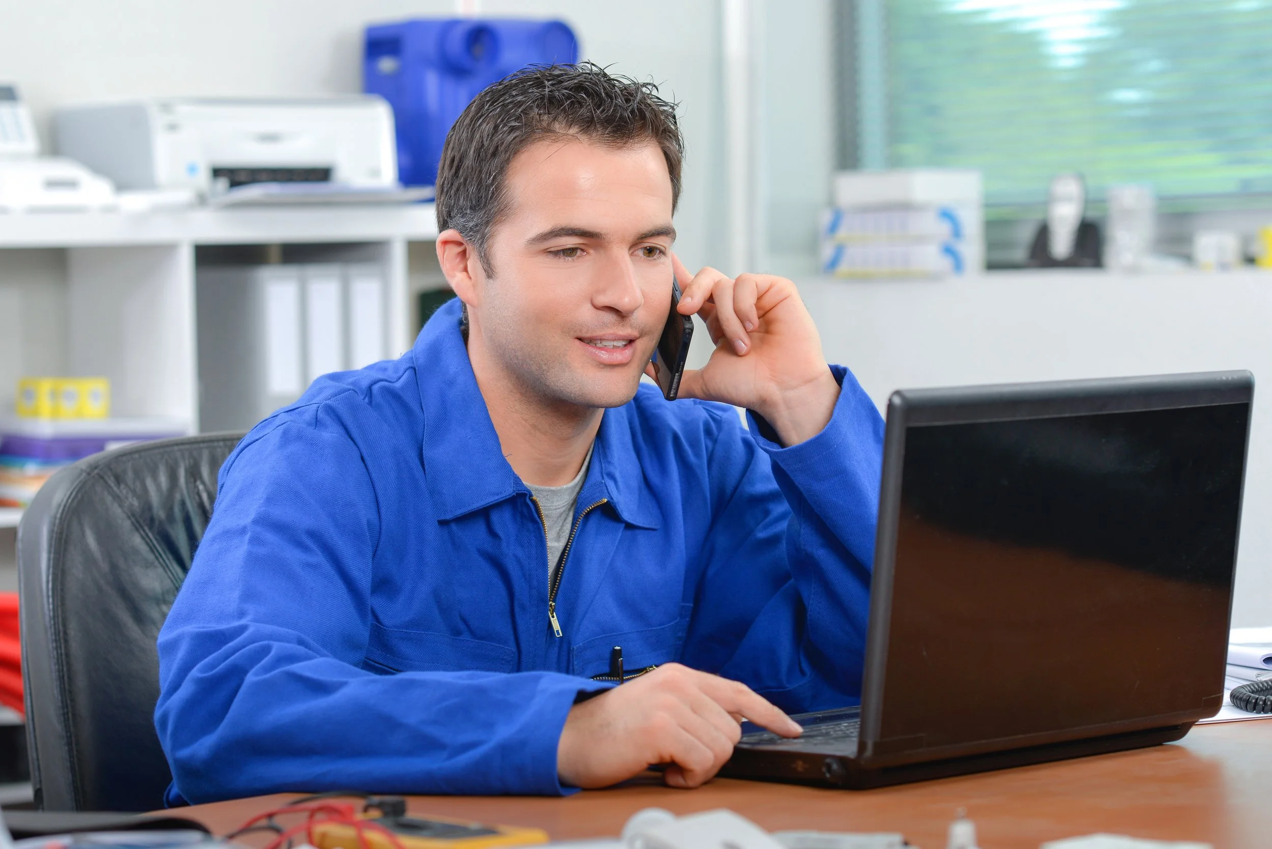 A man with short dark hair wearing a blue work jacket, sitting at a desk, speaking on a mobile phone, working on a laptop in an office with shelves and office equipment in the background.