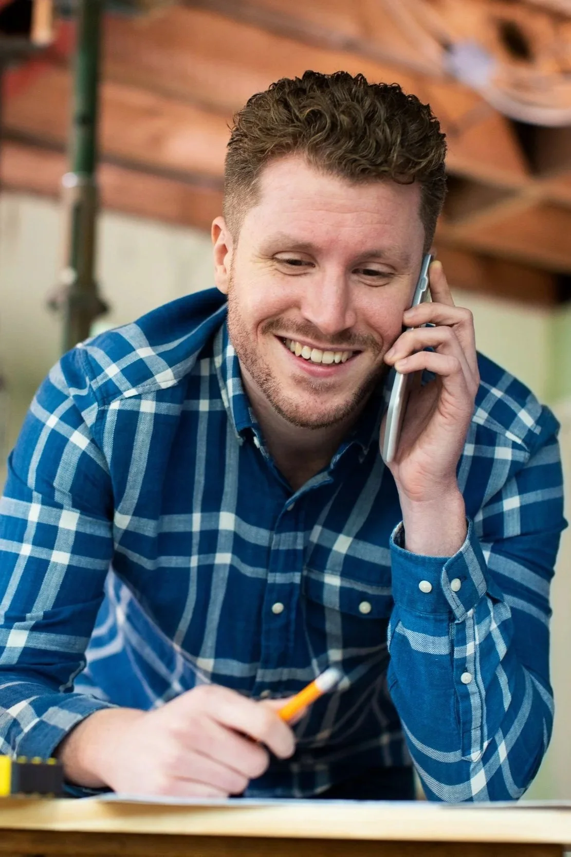 A man with curly hair and a beard, smiling and talking on a cellphone, wearing a blue plaid shirt, sitting at a table with a pen in hand, in a cozy indoor space.