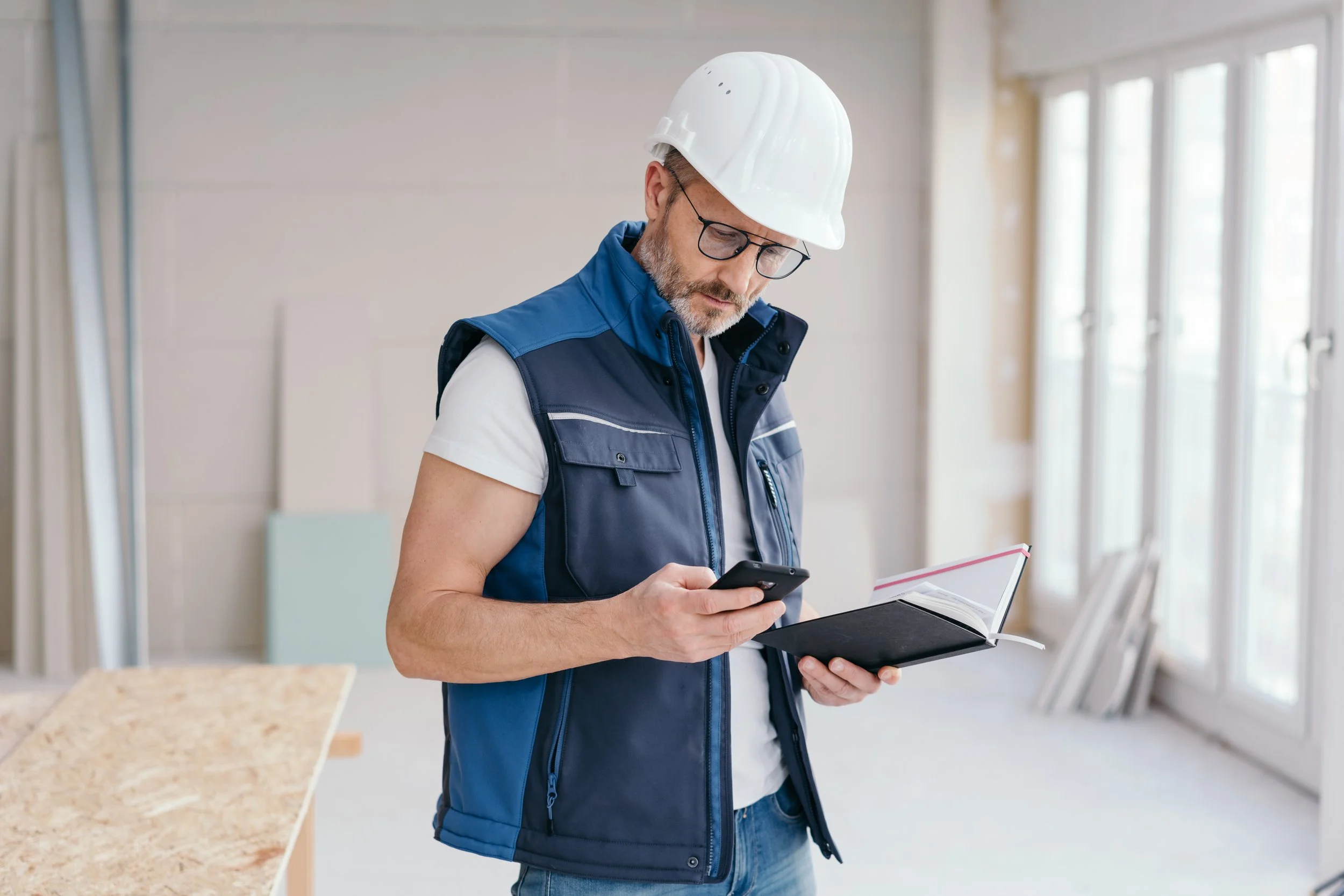 A man wearing a white hard hat, glasses, and a blue vest, standing in a construction or renovation site, looking at his smartphone while holding an open planner or notebook.