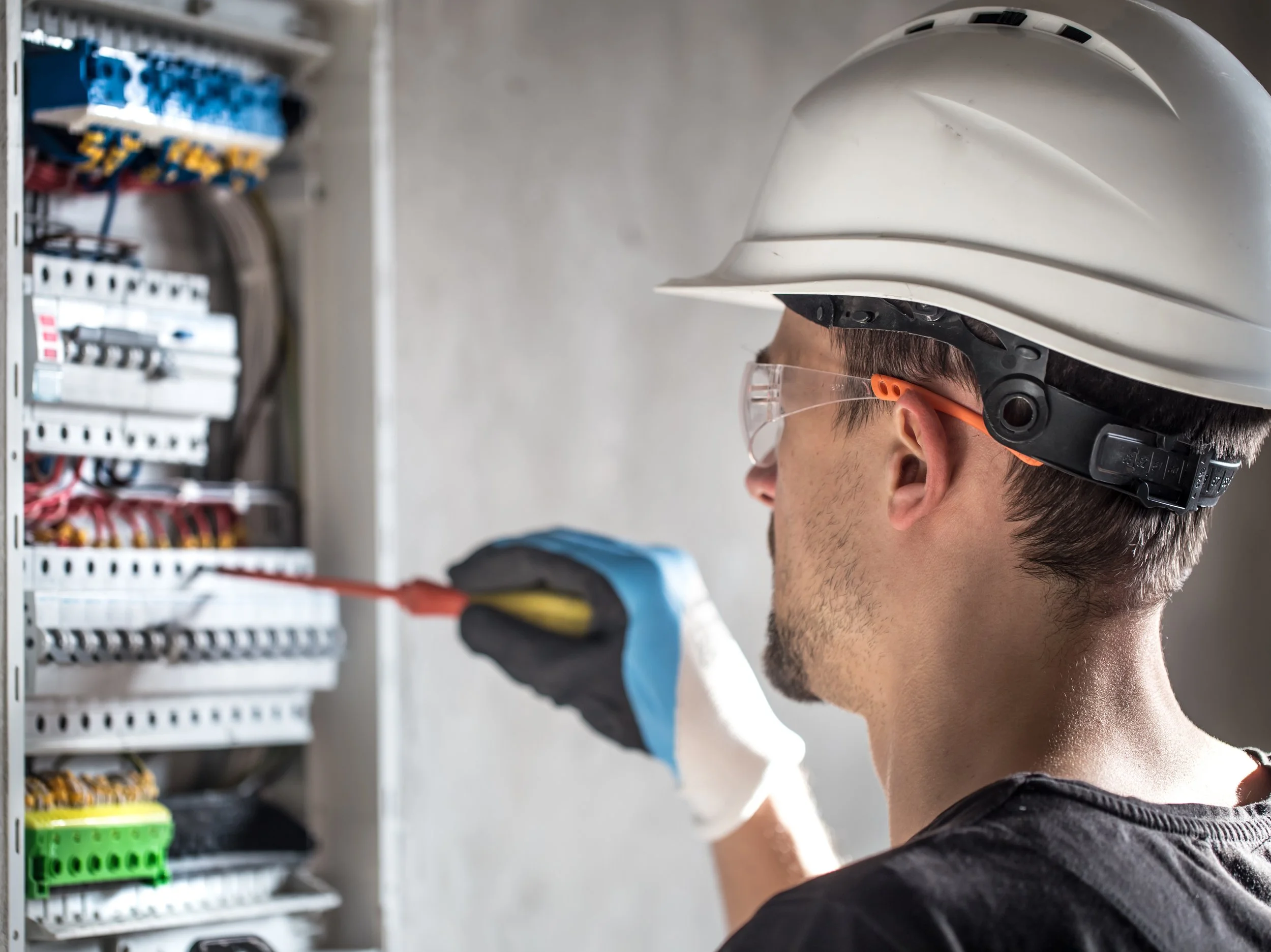 A man wearing safety glasses and a white hard hat working in an electrical or mechanical setting, with control panels or electrical components visible in the background.