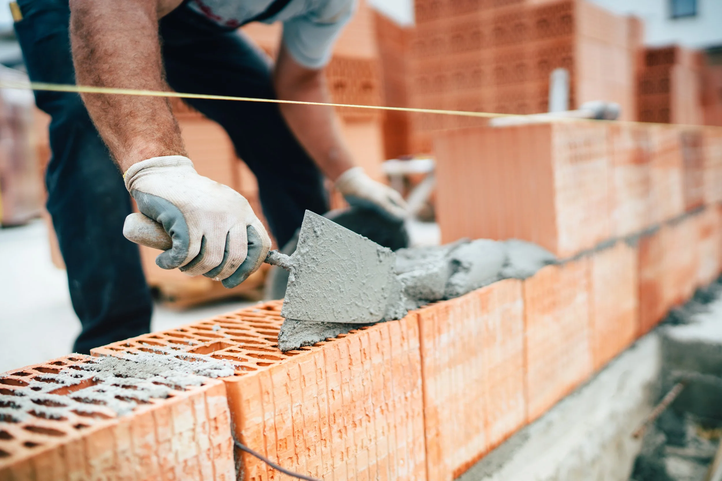 A construction worker wearing gloves smoothing wet cement onto a brick wall with a trowel, with stacks of bricks in the background.