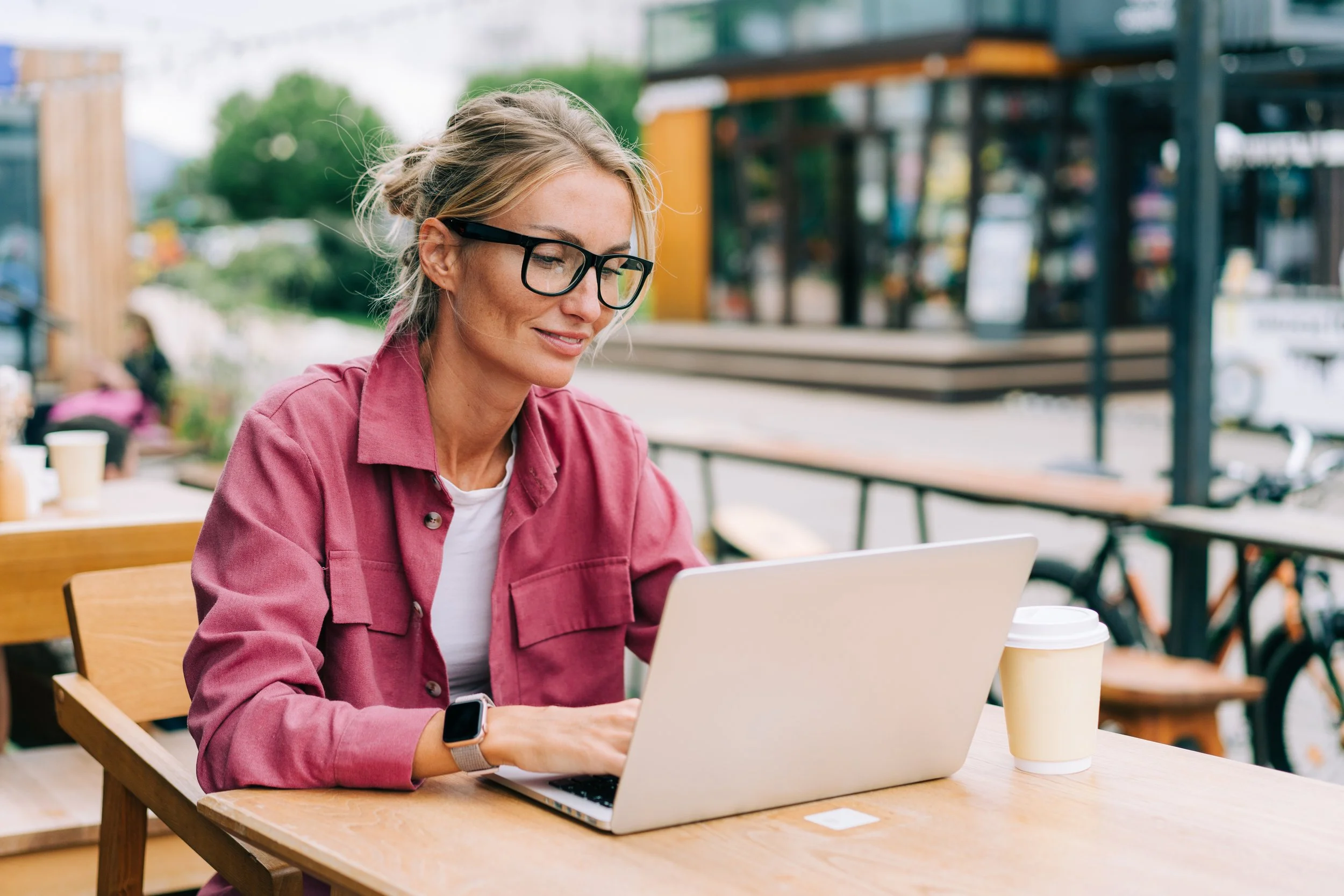 A woman with blonde hair, glasses, and wearing a pink jacket, sitting at an outdoor wooden table using a silver laptop, with a disposable coffee cup nearby, in a casual outdoor cafe or park setting.