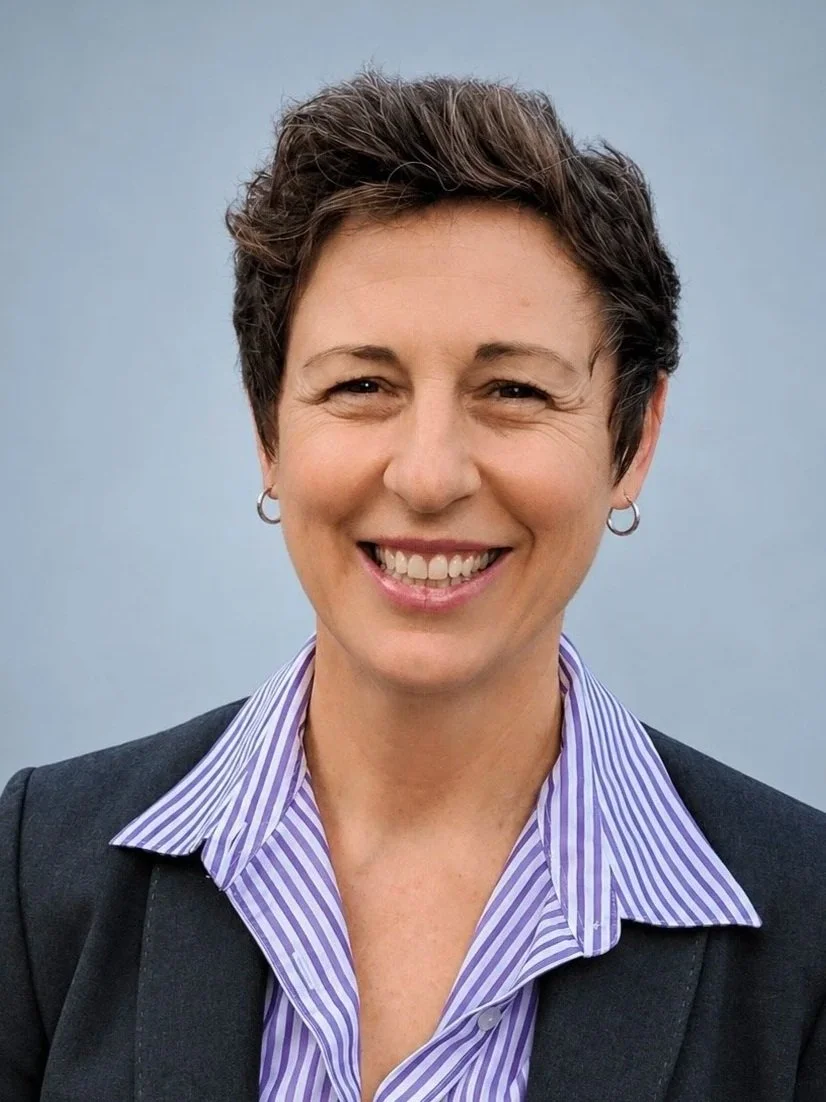 A woman, who is a member of the Clube Pickleball Lisboa Board, with short brown hair, smiling, wearing a striped blue and white collared shirt, a dark blazer, and hoop earrings, standing against a plain gray background.