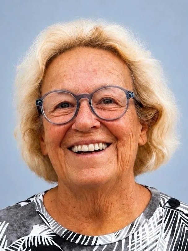 Close-up of an elderly woman, who is a member of the Clube Pickleball Lisboa Board, with blonde hair, wearing glasses, smiling, and dressed in a black and white patterned top against a light blue background.