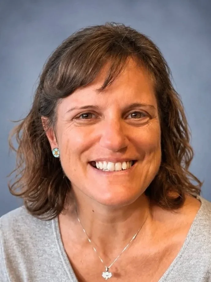 Close-up of a smiling woman, who is a member of the Clube Pickleball Lisboa Board, with shoulder-length brown hair, wearing turquoise earrings, a silver necklace with a butterfly pendant, and a light gray top against a blue background.