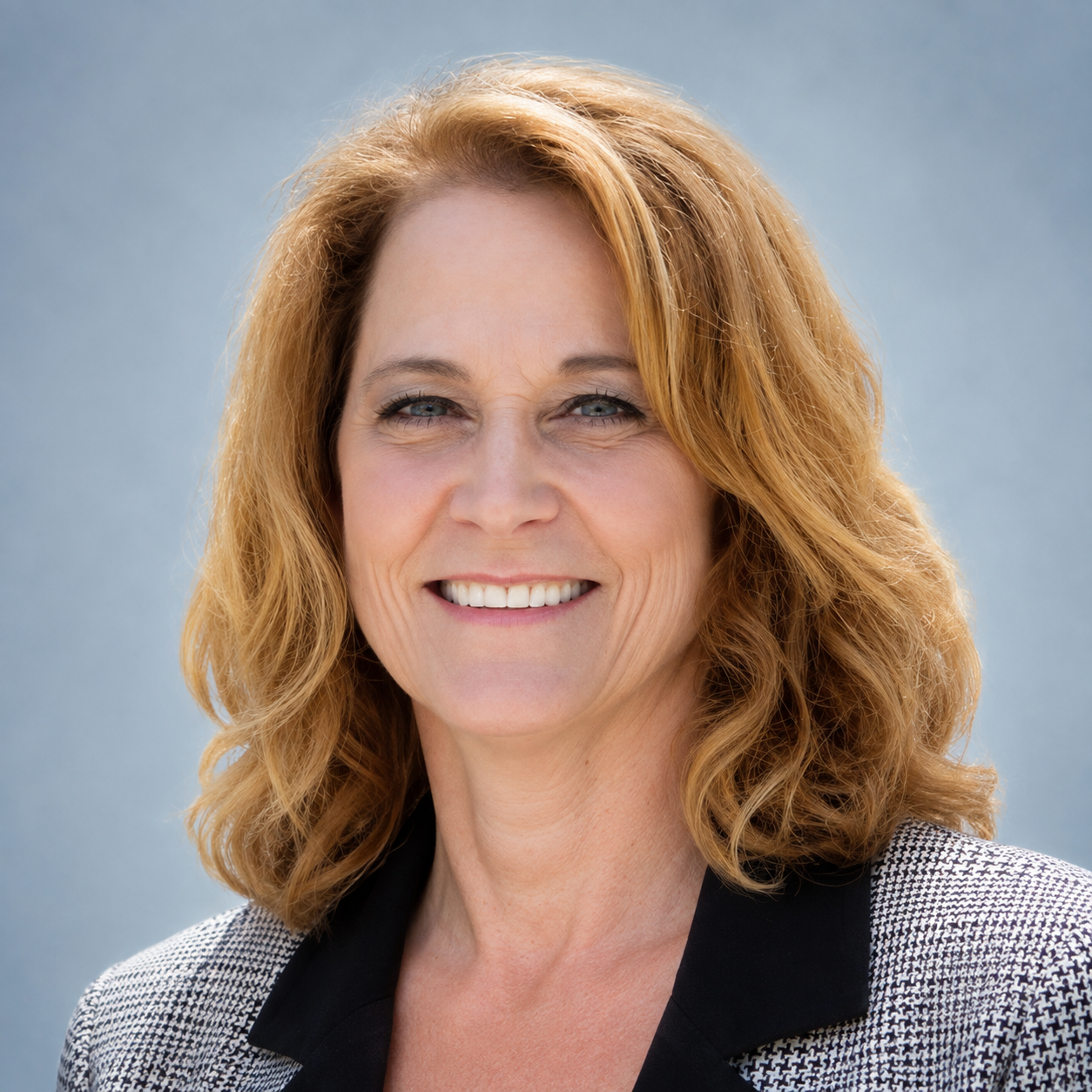 Close-up portrait of a smiling middle-aged woman, who is a member of the Clube Pickleball Lisboa Board, with shoulder-length wavy blonde hair wearing a black top and a checkered blazer against a light blue background.