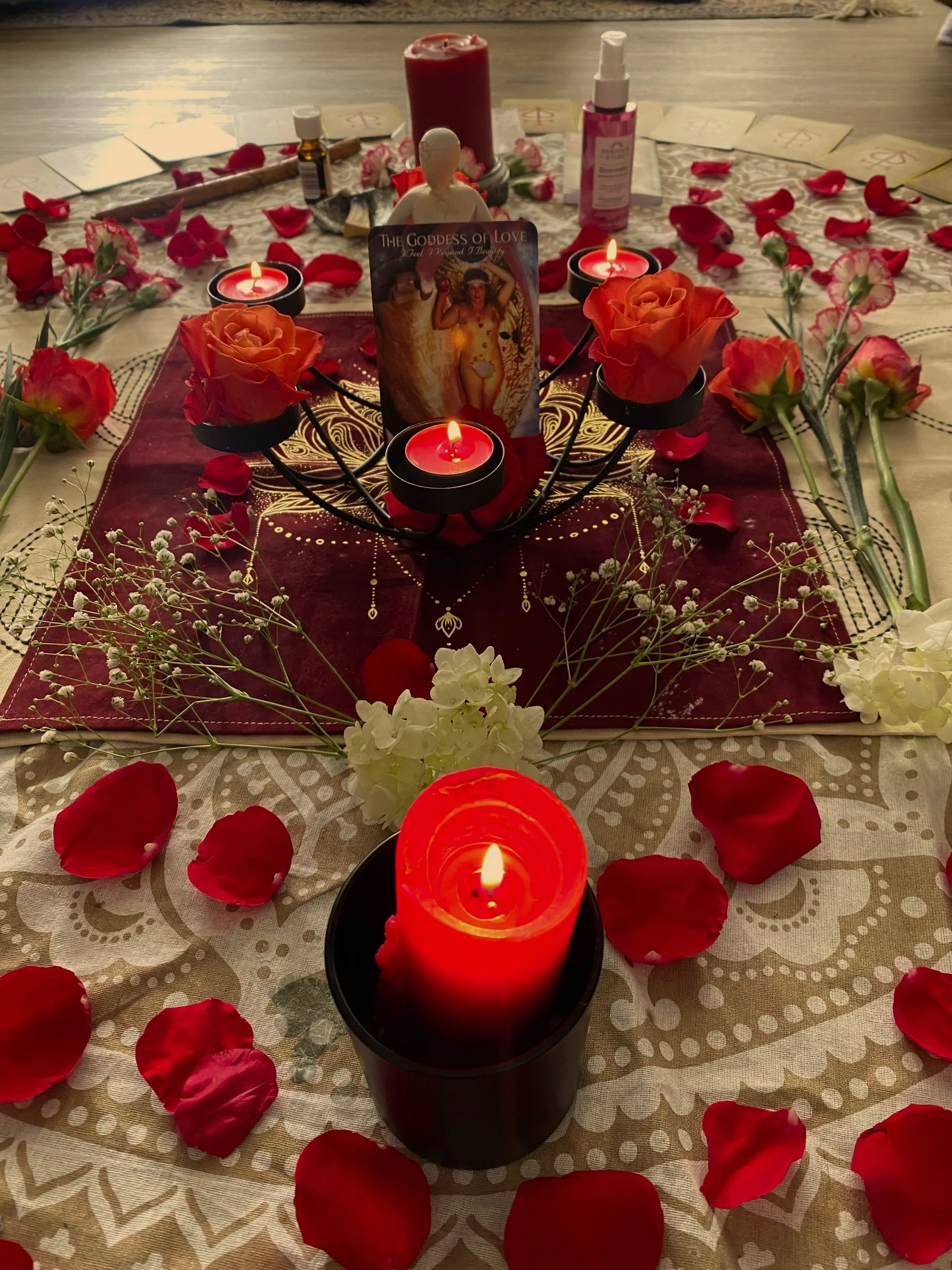 A romantic altar decorated with red candles, fresh roses, rose petals, white flowers, a goddess of love card, and an angel figurine, set on a table with a red and white patterned cloth for a special occasion.
