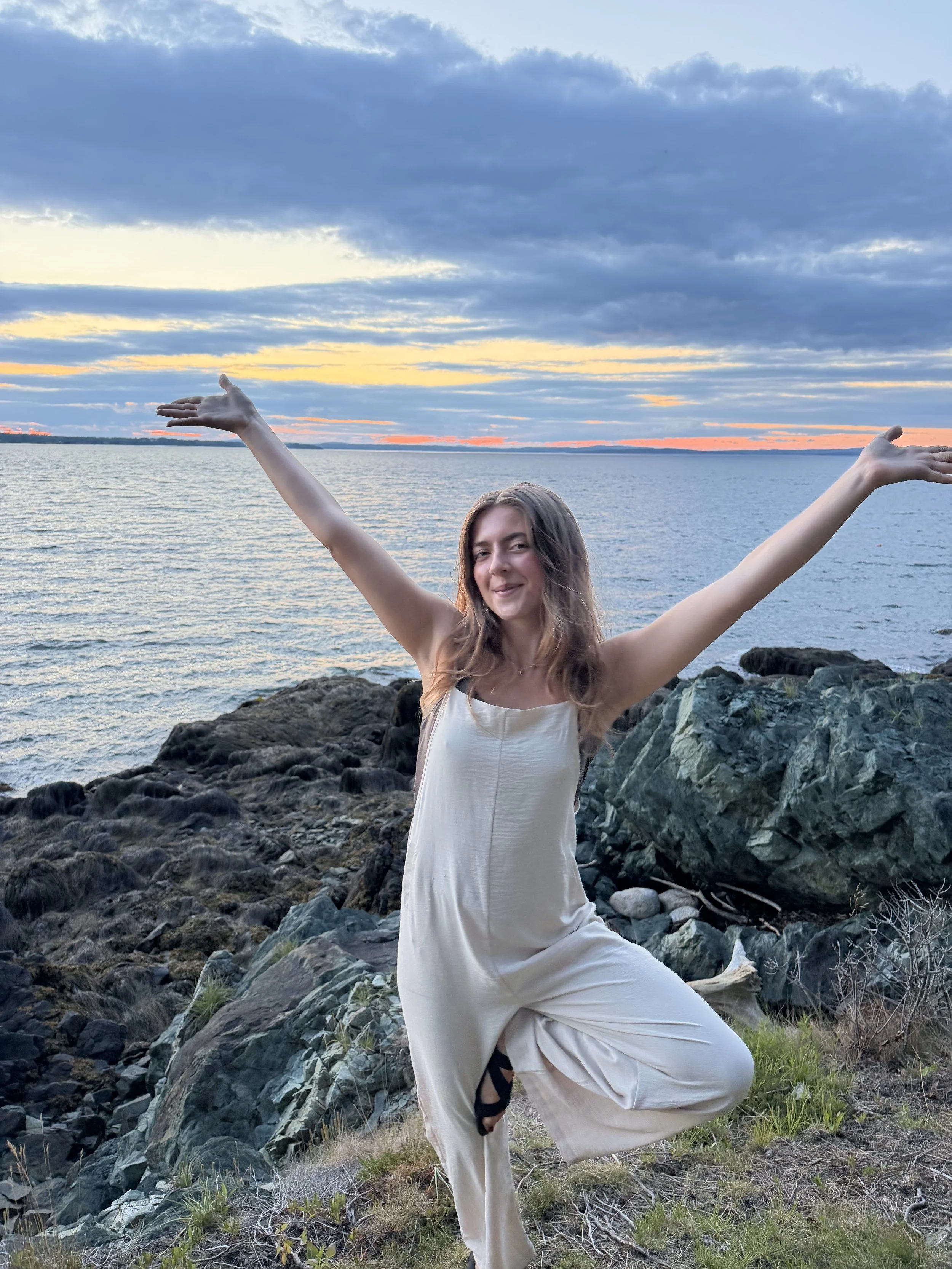 A woman with long hair, dressed in a white dress, practicing yoga outdoors on rocky terrain near a body of water during sunset with a sky filled with clouds.