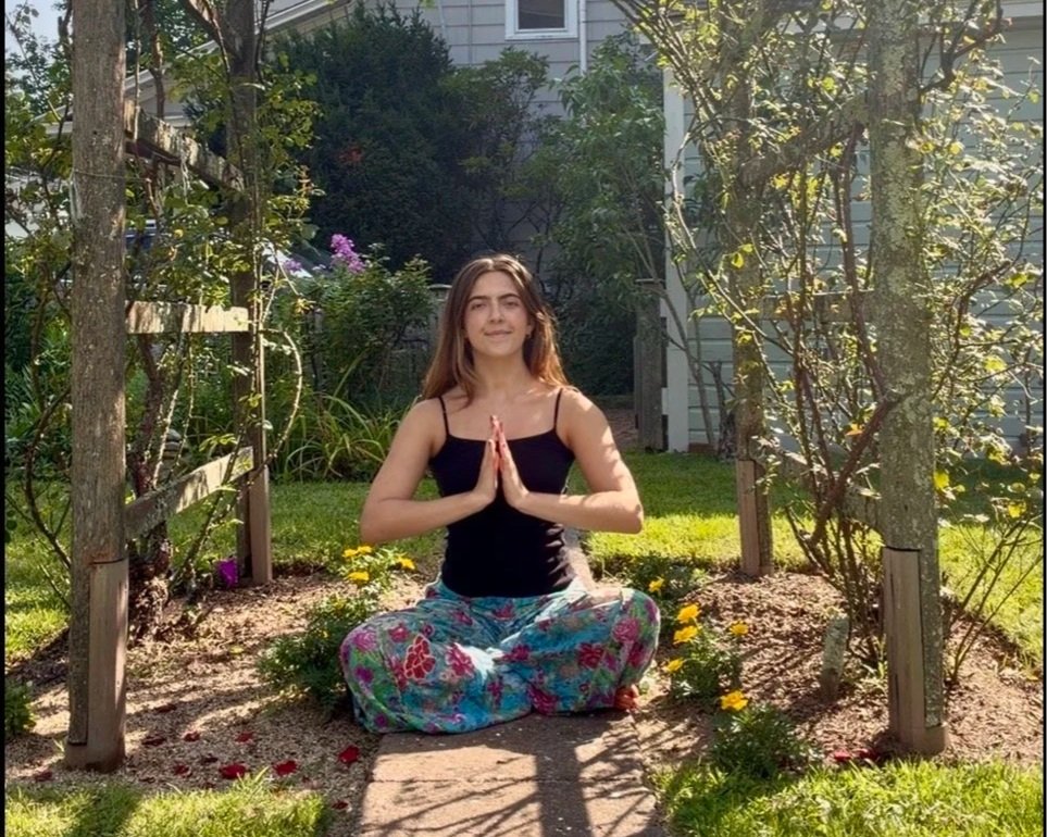 A young woman sitting in a garden with her legs crossed in a meditative pose, hands together in a prayer position, smiling with eyes closed, surrounded by yellow flowers, trees, and greenery, with sunlight shining.