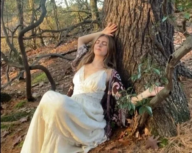 A woman in a white dress sitting against a large tree in a wooded area, with her eyes closed and one hand resting on her forehead, appearing peaceful and relaxed.