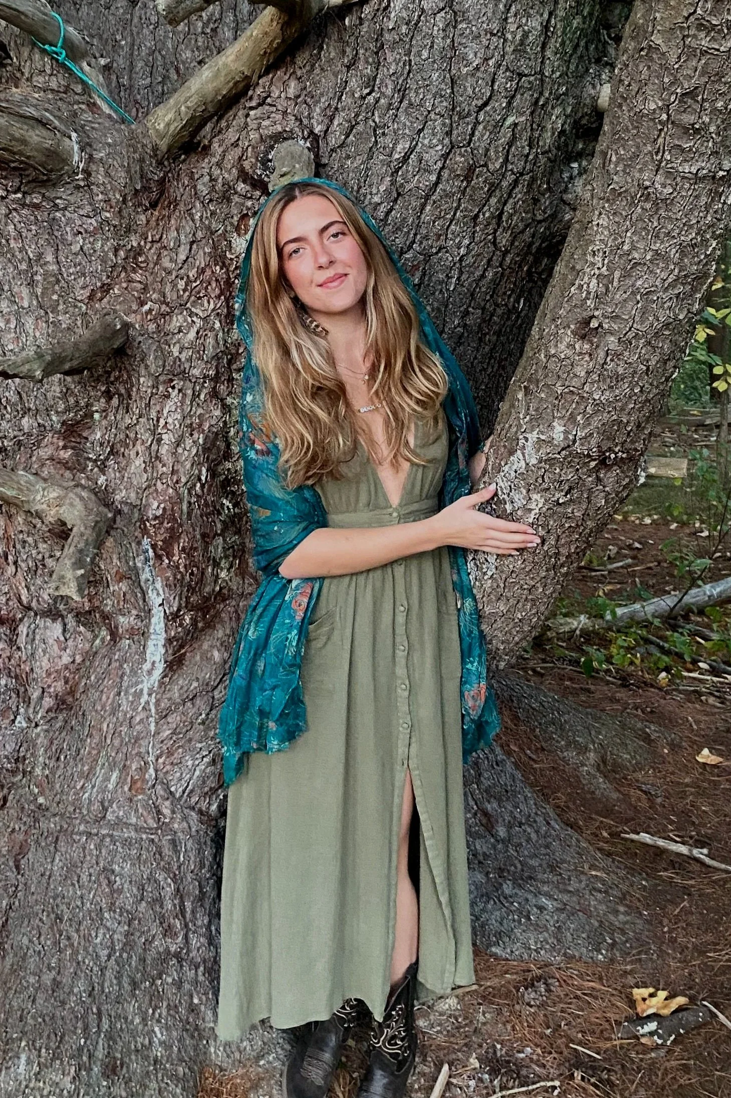 A woman standing in front of a large tree, wearing a green dress, black boots, and a blue floral shawl, with her eyes slightly closed and smiling.