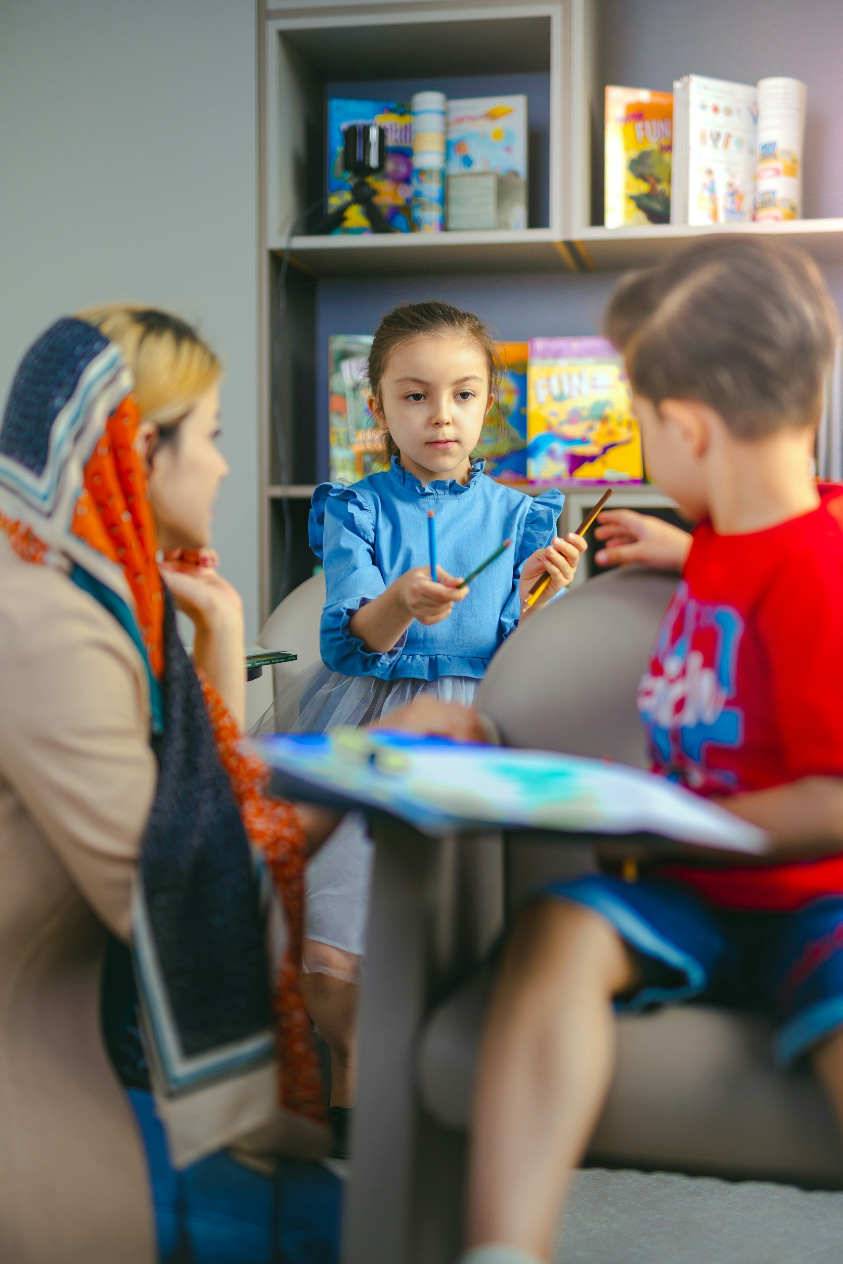 A group of children sitting around a table in a classroom, engaging in a discussion or activity. One girl in a blue dress is holding colored pencils, and there are books and games on the shelves behind them.